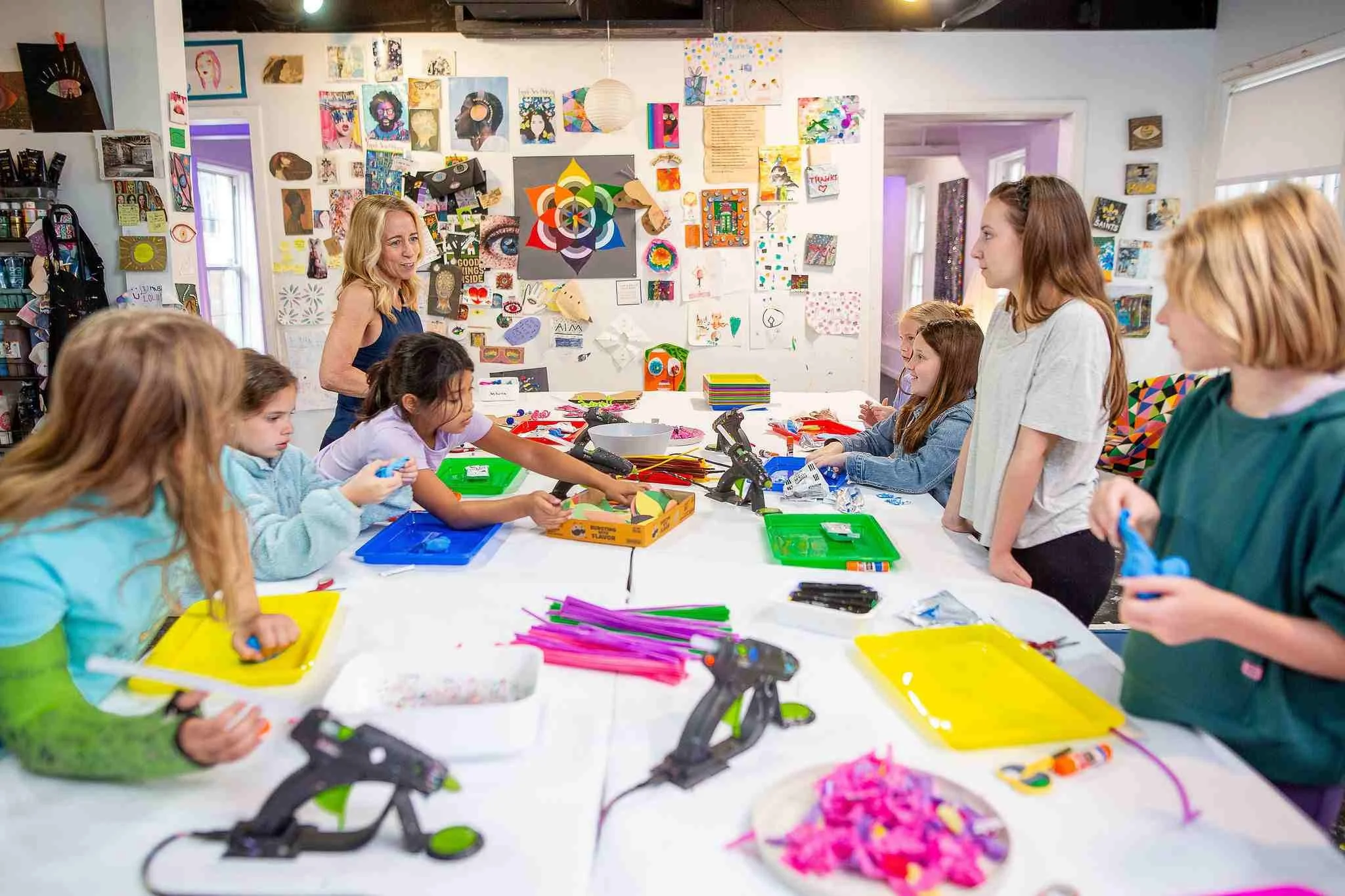 A group of children and a woman in an art studio, engaging in arts and crafts activities at a long table filled with colorful supplies. The walls are decorated with various artworks.