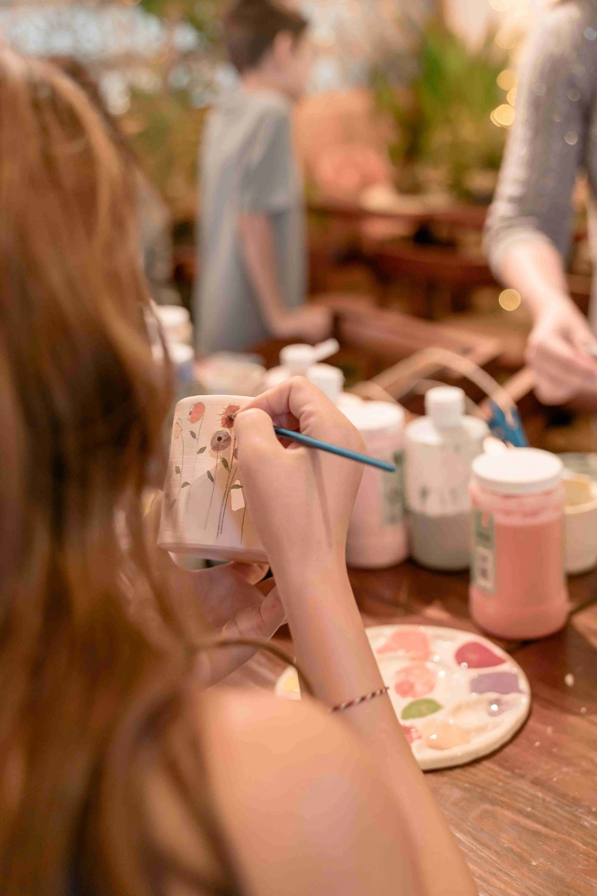 Close-up of a person painting on a ceramic mug with floral patterns, with art supplies and a palette of paint on a wooden table in the background, and other people in a blurred setting.