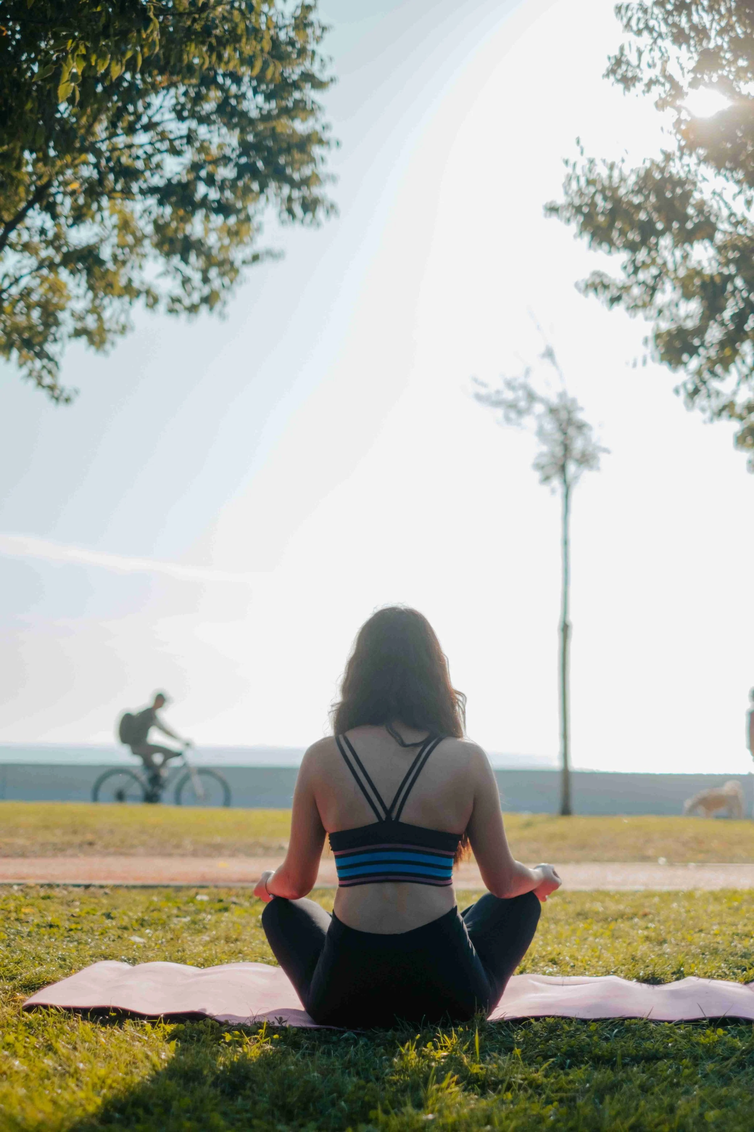 A woman practicing yoga outdoors on a pink mat, sitting cross-legged with her back to the camera. In the background, there are trees, a person riding a bicycle, and the sun shining through the leaves.