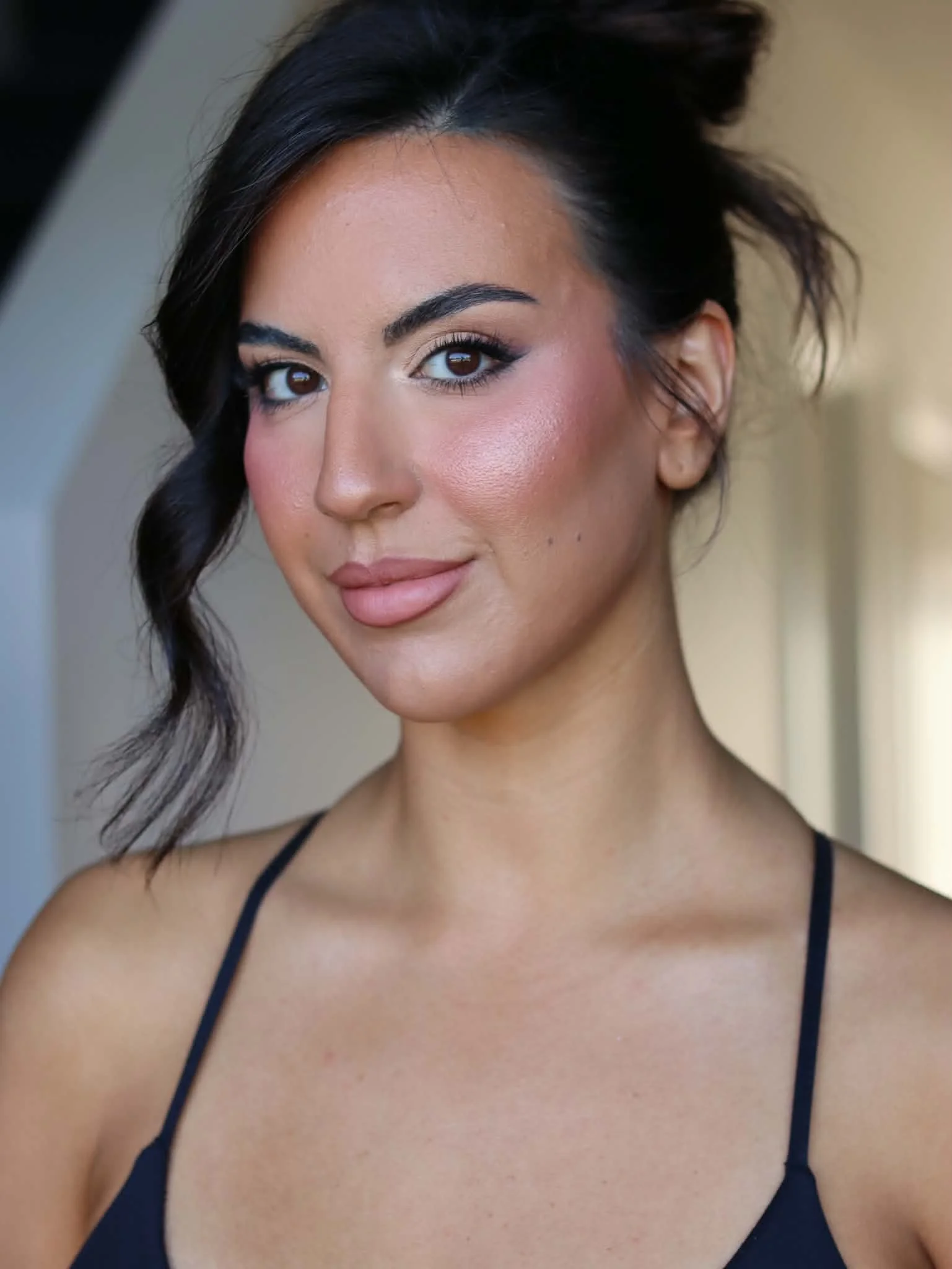 A young woman with dark hair styled in loose curls, tan skin, and wearing a black spaghetti strap top, smiling subtly in a close-up portrait.