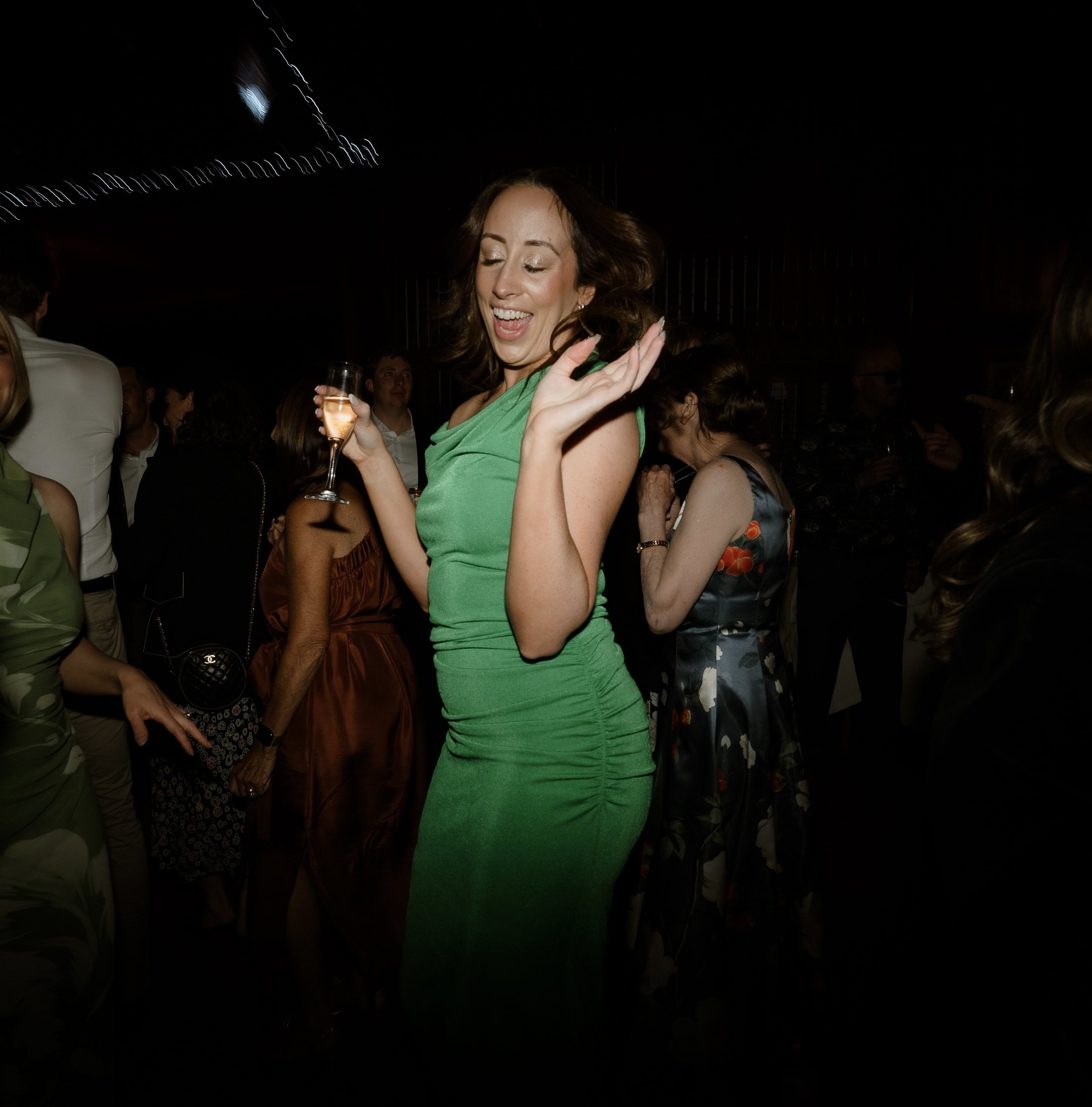 A woman in a green dress dancing and smiling at a party at night, holding a glass of champagne, surrounded by other people.