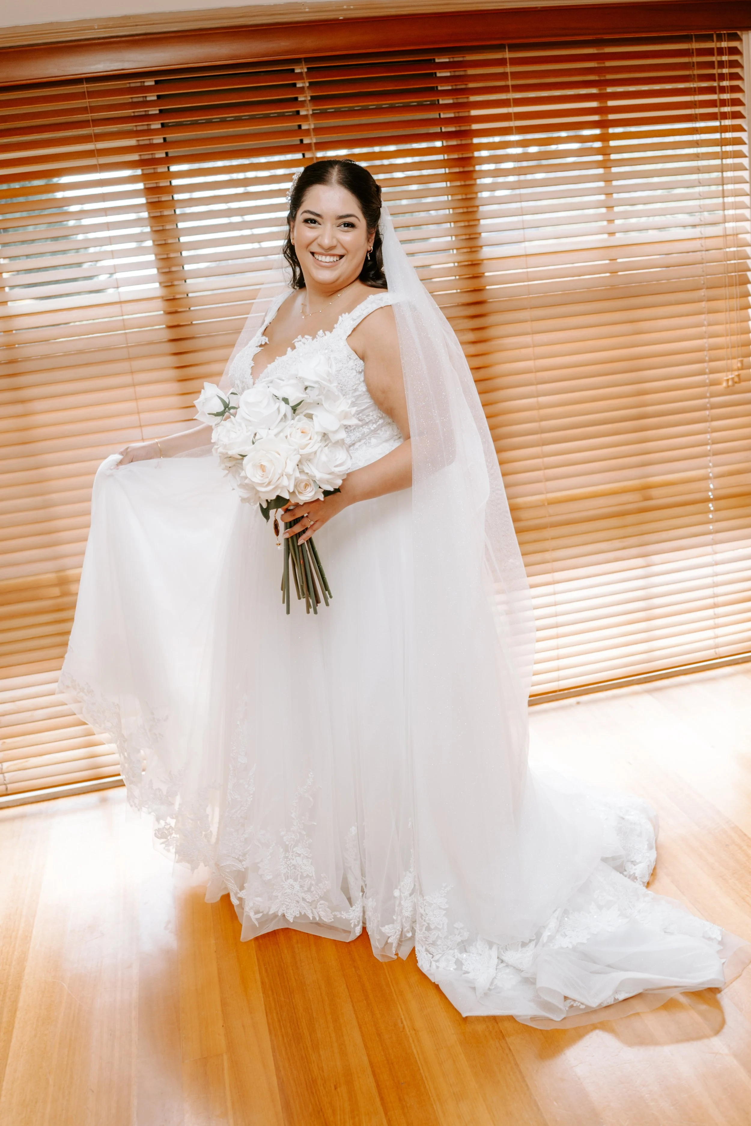 A bride in a white wedding gown holding a bouquet of white roses and smiling. She is standing indoors in front of wooden blinds with sunlight filtering through.
