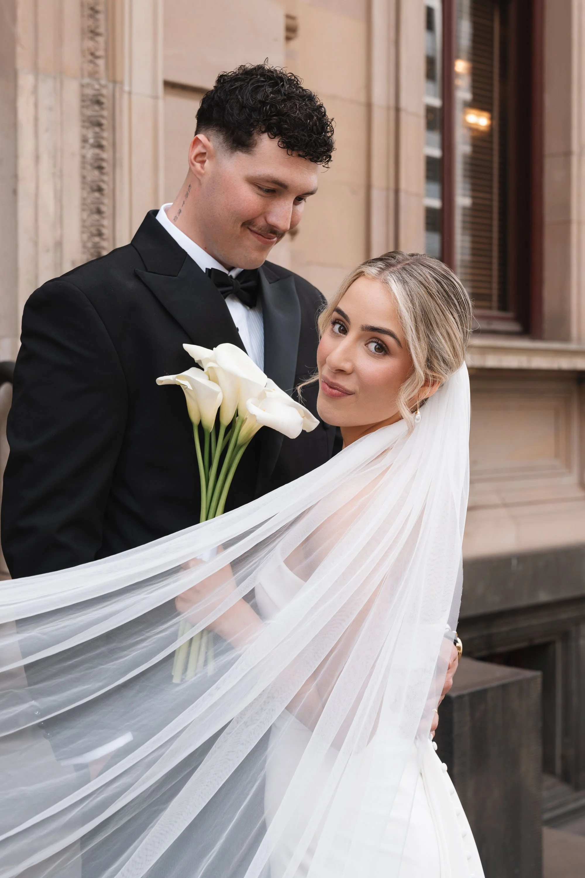 A newlywed couple standing in front of a building, with the groom holding a bouquet of white calla lilies, both dressed in wedding attire, smiling and looking at each other.