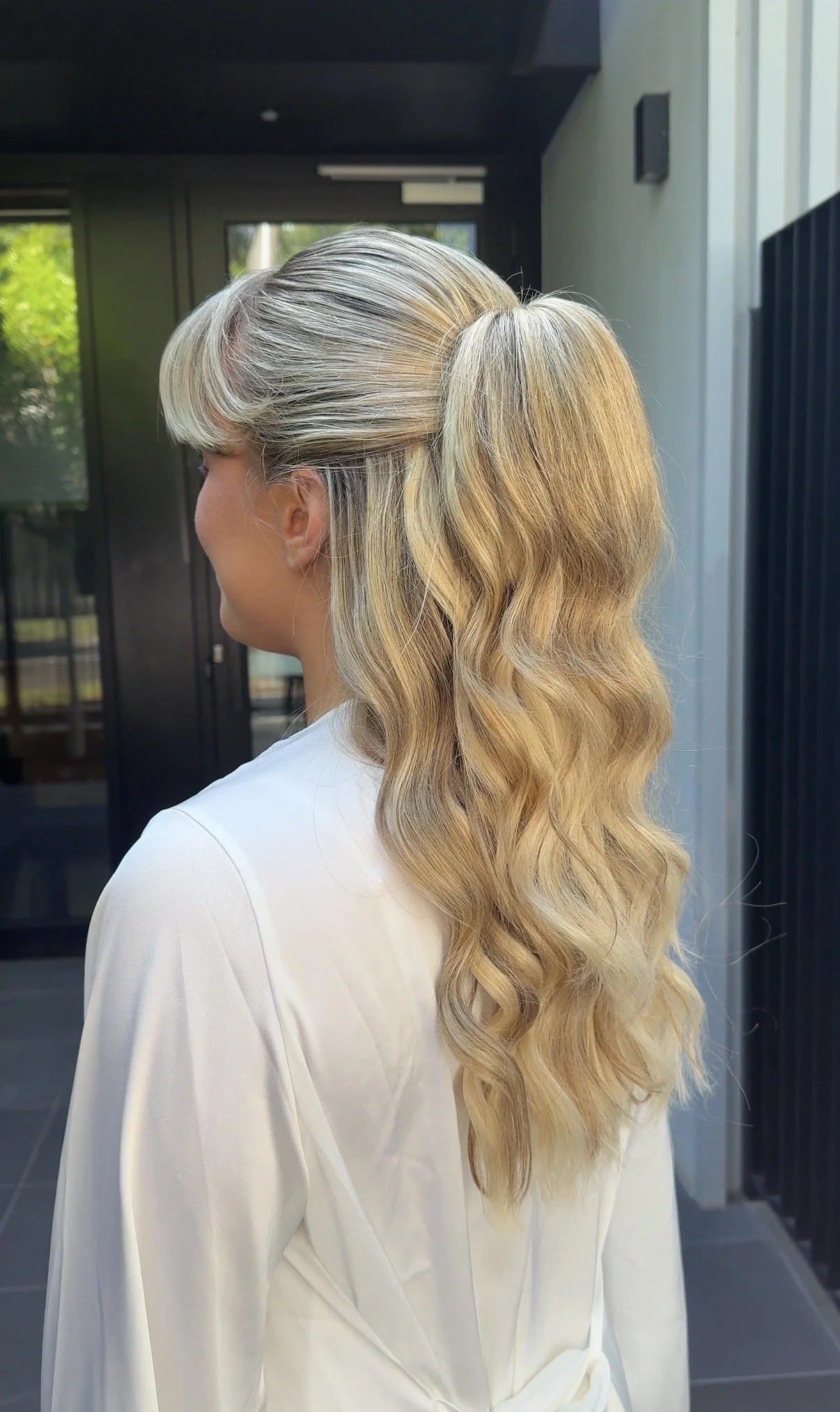 A woman with long, wavy blonde hair styled in loose waves, wearing a white blouse, standing outside near a modern building.