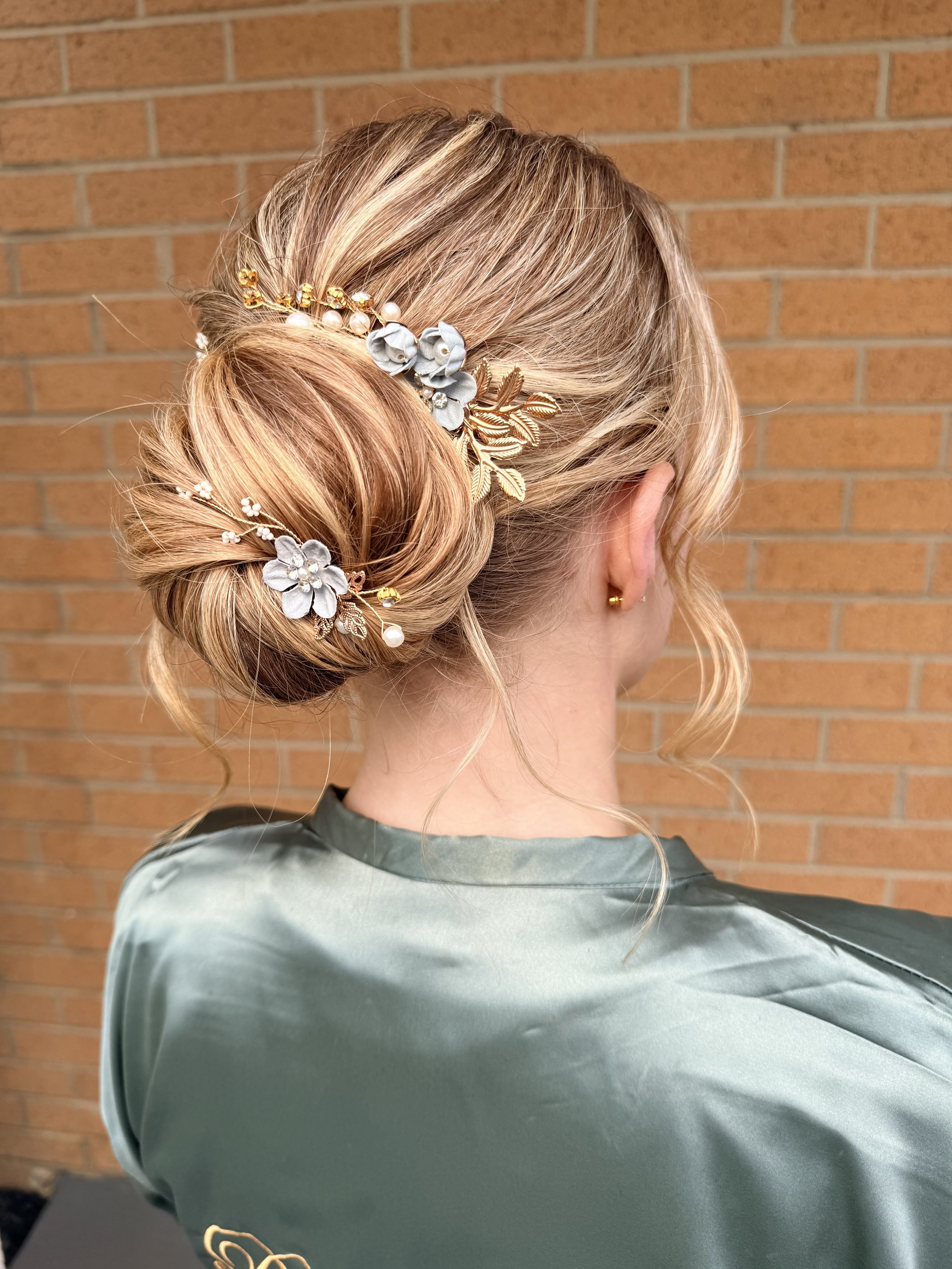 A woman with styled blonde hair with curls, wearing a floral and pearl headpiece, wearing a shiny satin or silk green robe, standing in front of a brick wall.
