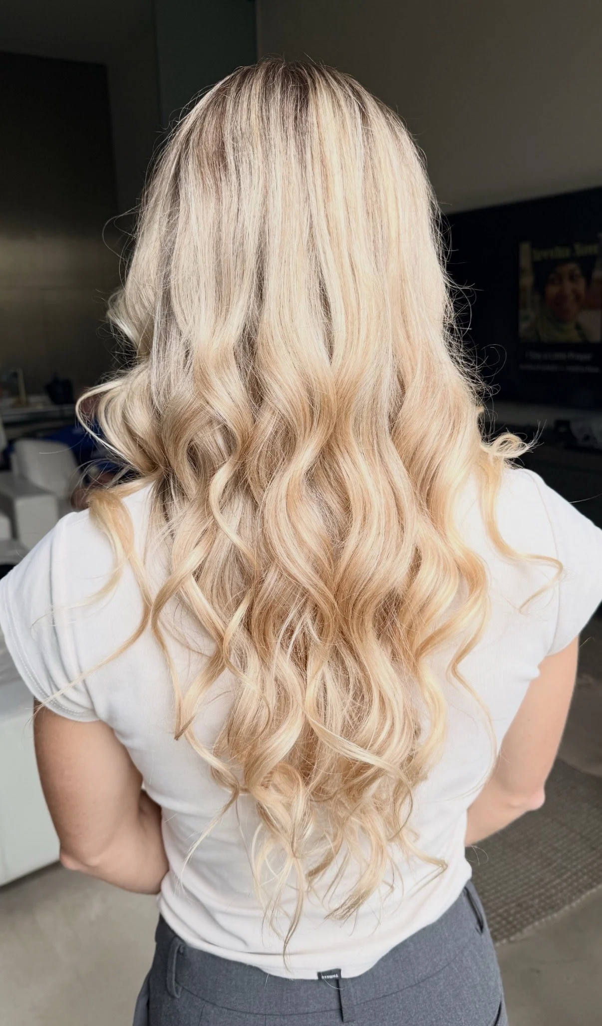 Back view of woman with long, wavy, blonde hair wearing a white t-shirt and gray pants, standing indoors.