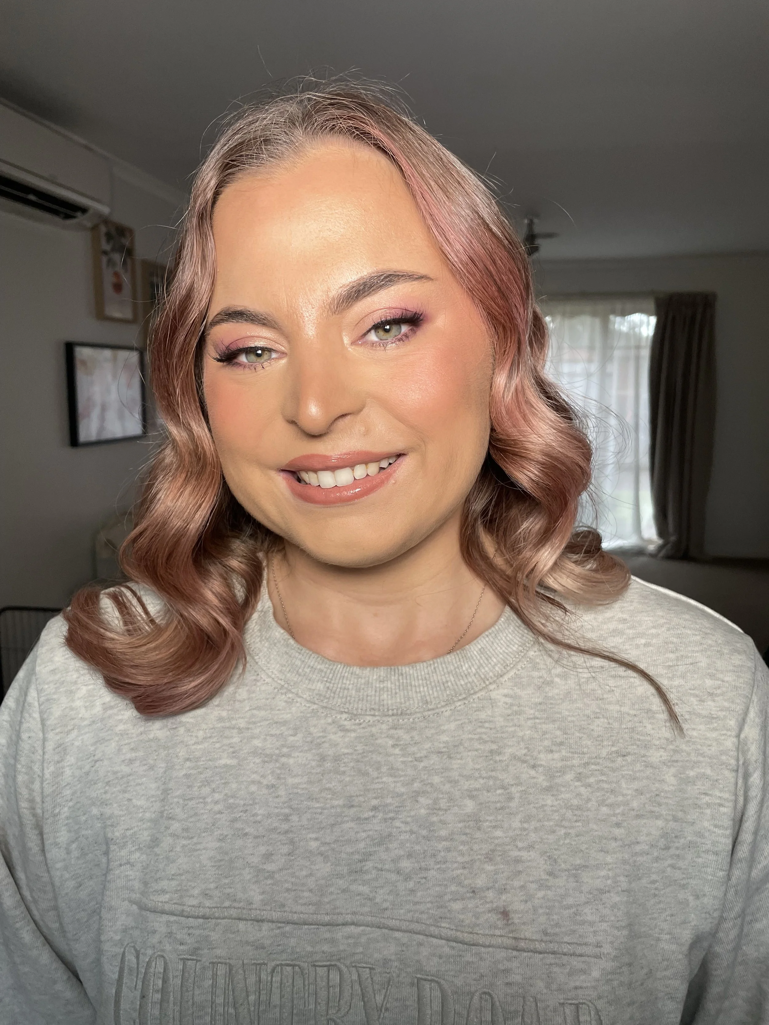 A woman with pink wavy hair, light makeup, and a light-colored sweatshirt smiling in a room with curtains and picture frames in the background.