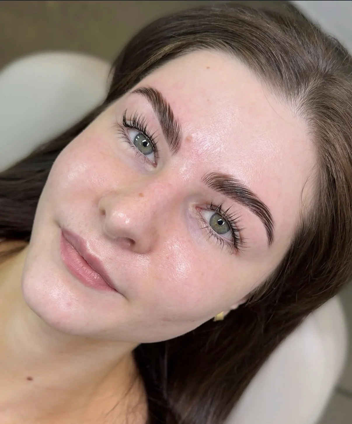 Close-up of a woman with fair skin, green eyes, long lashes, well-groomed eyebrows, and brown hair lying on a surface.