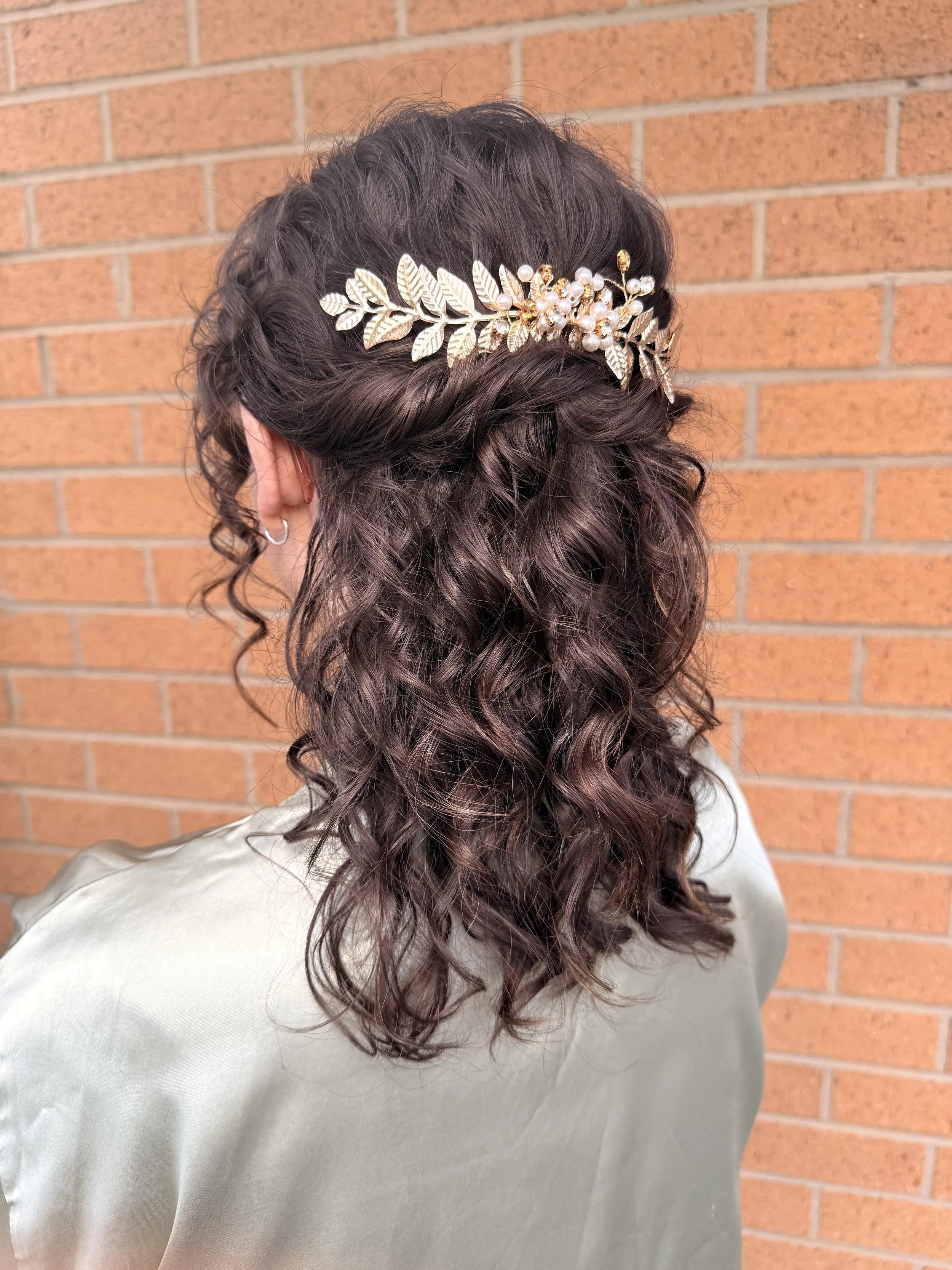 Back of woman with long, curly, dark brown hair wearing a decorative hair accessory with gold leaves and pearls, standing against a brick wall.