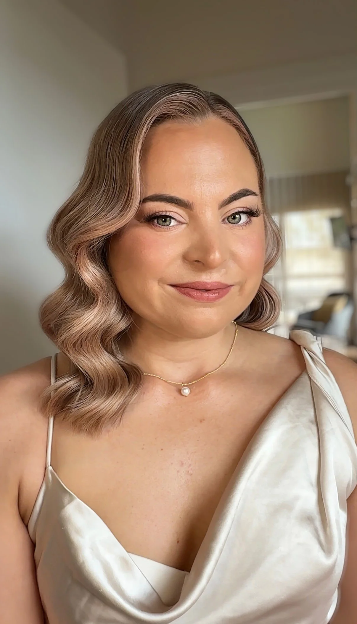 A woman with wavy, shoulder-length hair and light skin, wearing a satin cream-colored dress and a delicate pearl necklace, smiling in a well-lit room with beige walls and a window with curtains.