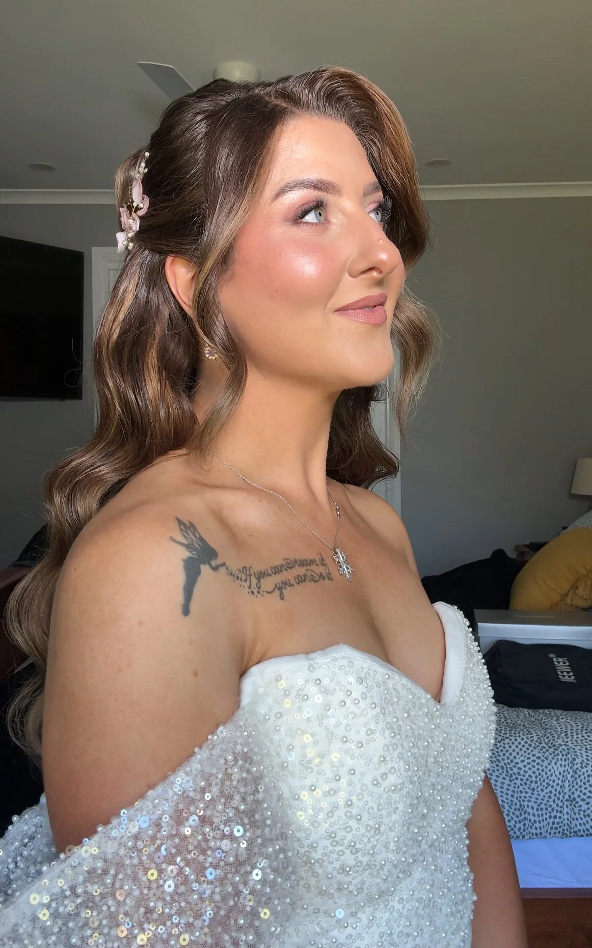 A woman in a white dress with sequins, wearing a necklace and earrings, with a tattoo on her shoulder, smiling and looking to the side indoors.
