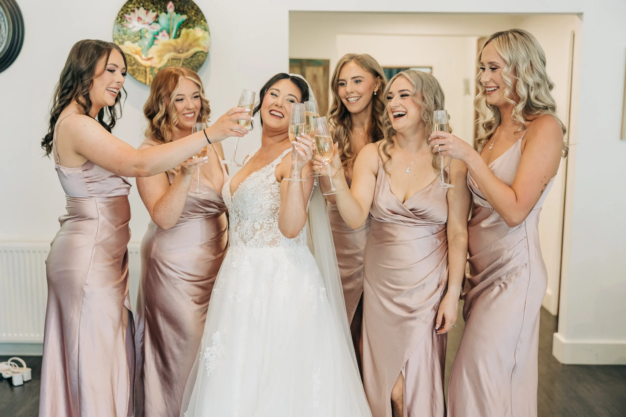 A bride in a white wedding gown surrounded by six bridesmaids in matching pink satin dresses, all smiling and holding champagne glasses in celebration.