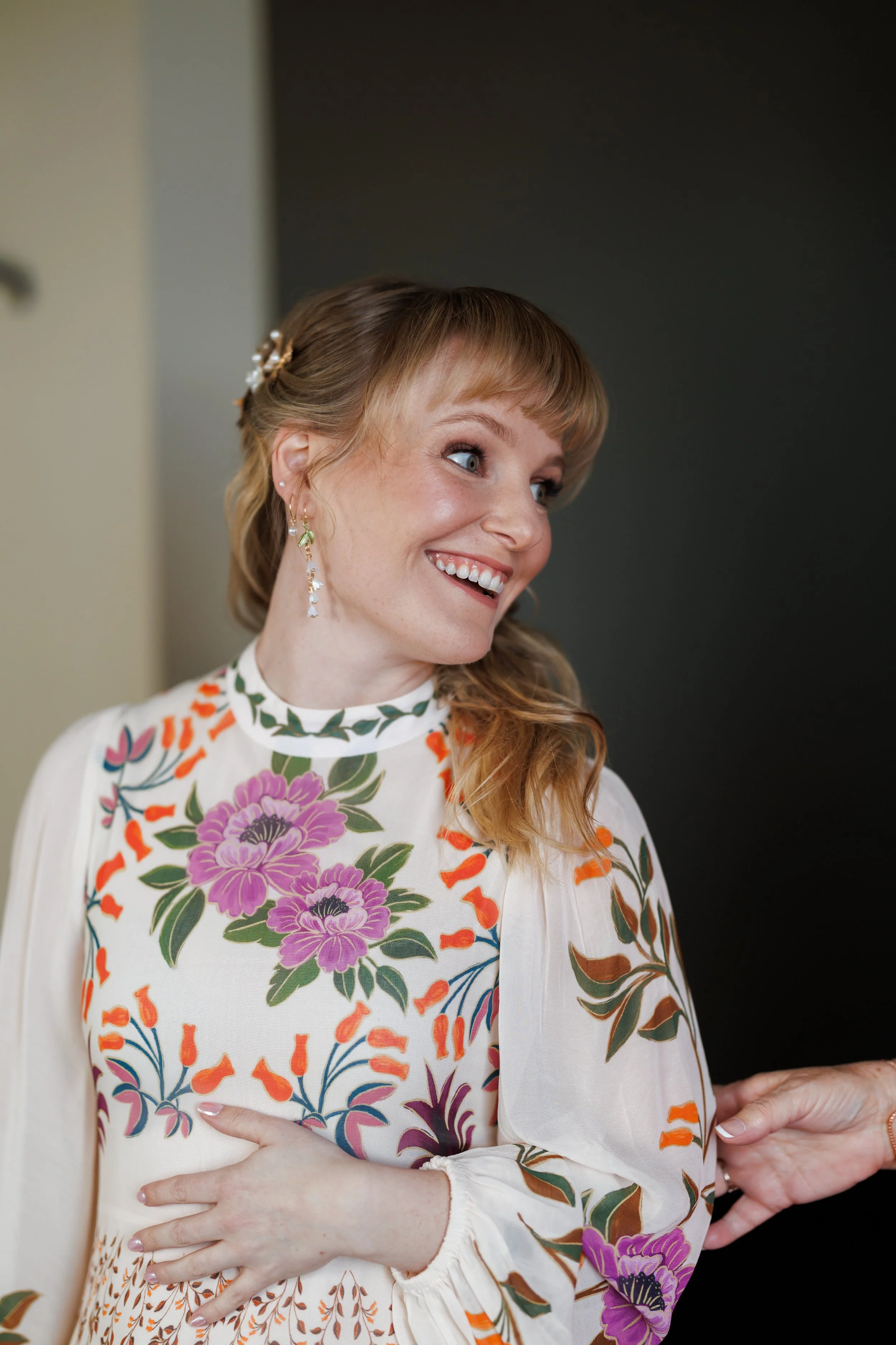 A woman with blonde hair styled with a pearl hair accessory and wearing floral earrings smiles while looking to her left. She is dressed in a white dress with colorful floral and orange accents, with her left hand resting on her stomach.