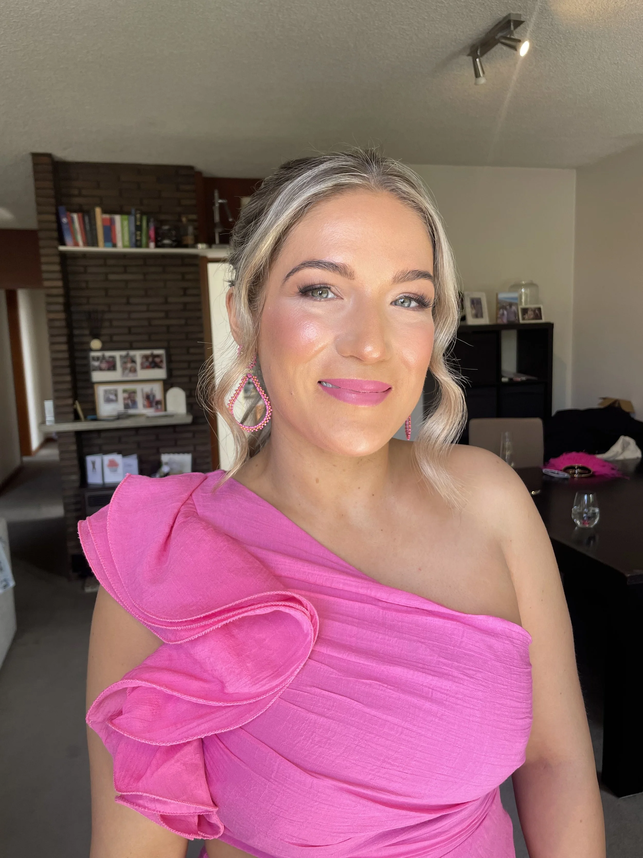 Close-up of a woman with blonde hair styled in loose curls, wearing pink earrings and a pink dress with ruffled off-shoulder sleeves, smiling softly in a well-lit room.
