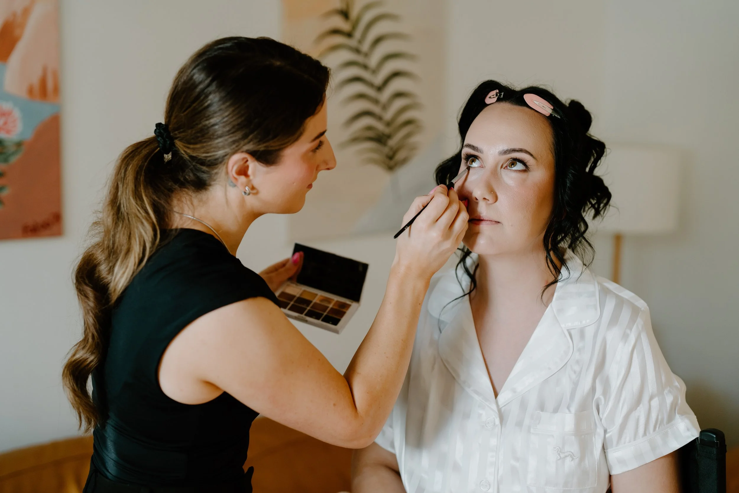 A makeup artist applies makeup under the eye of a woman with black curly hair, pink hair clips, and a white striped shirt, in a room with minimal decor.