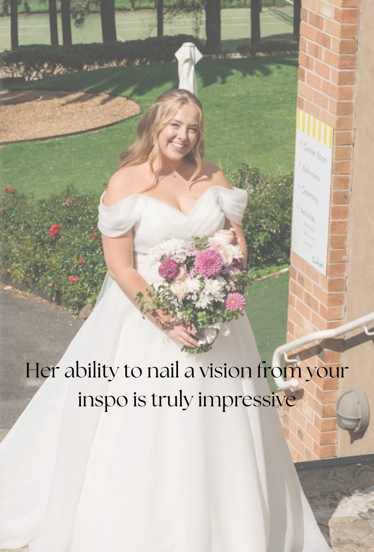 A smiling woman in a white off-shoulder wedding dress holding a bouquet of pink and white flowers outside on a sunny day near a building with a brick wall. There is a signboard and garden in the background.