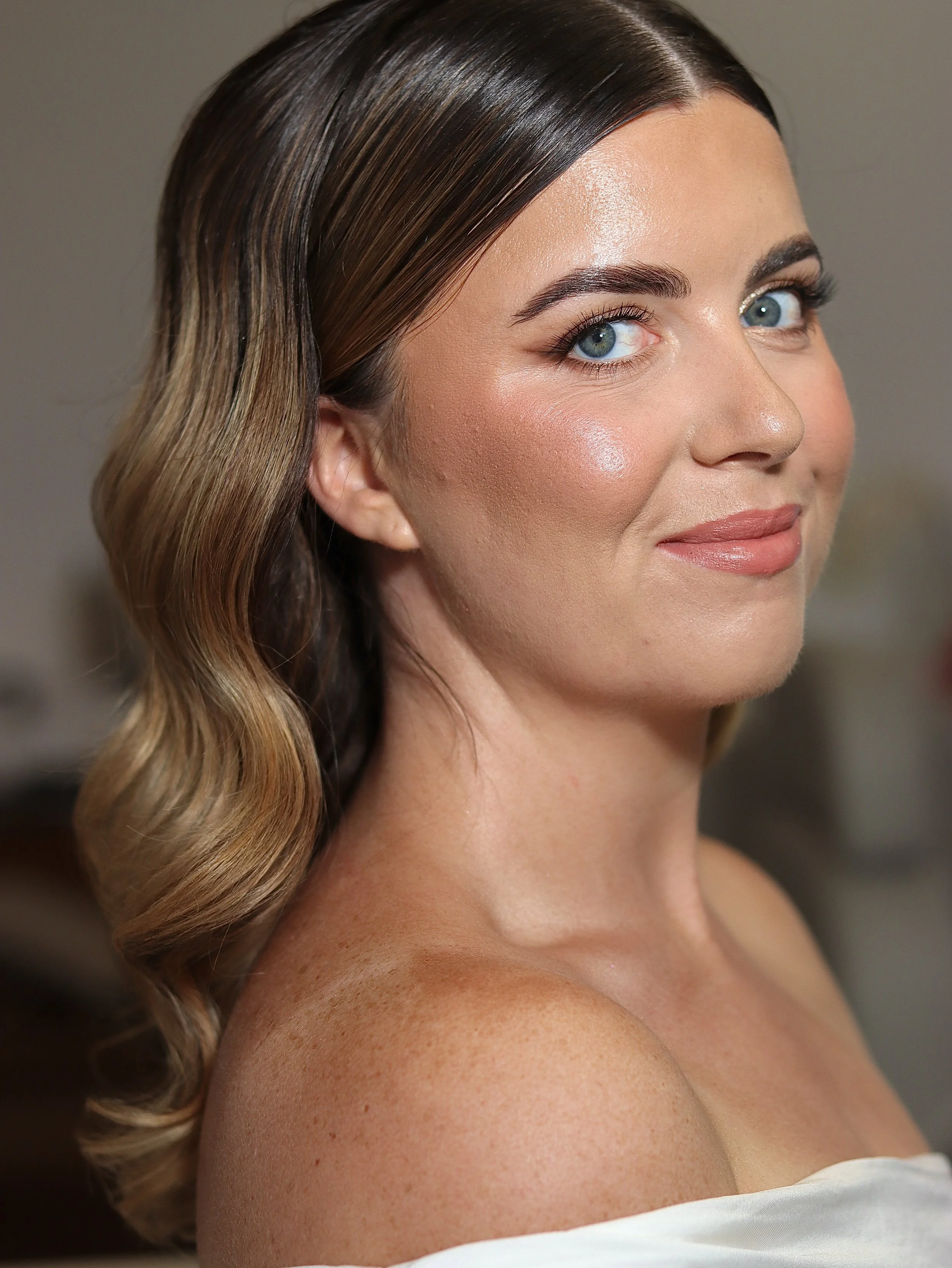 Close-up of a smiling woman with blue eyes, wearing makeup, with wavy brown hair, and wearing a white top.