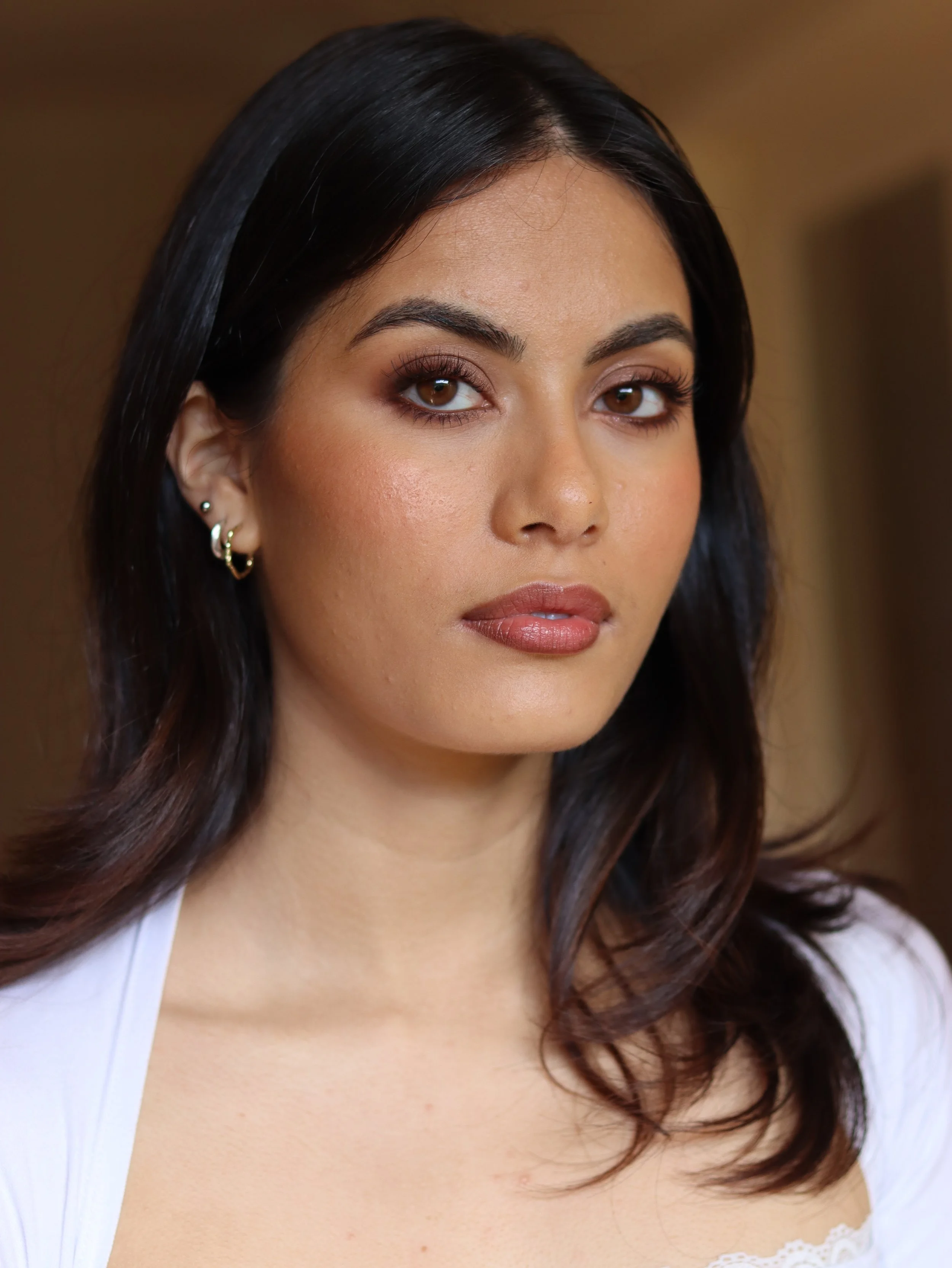A woman with dark, wavy hair, wearing a white top with lace at the neckline, facing slightly to the side, with makeup including eyeshadow, mascara, and lipstick.
