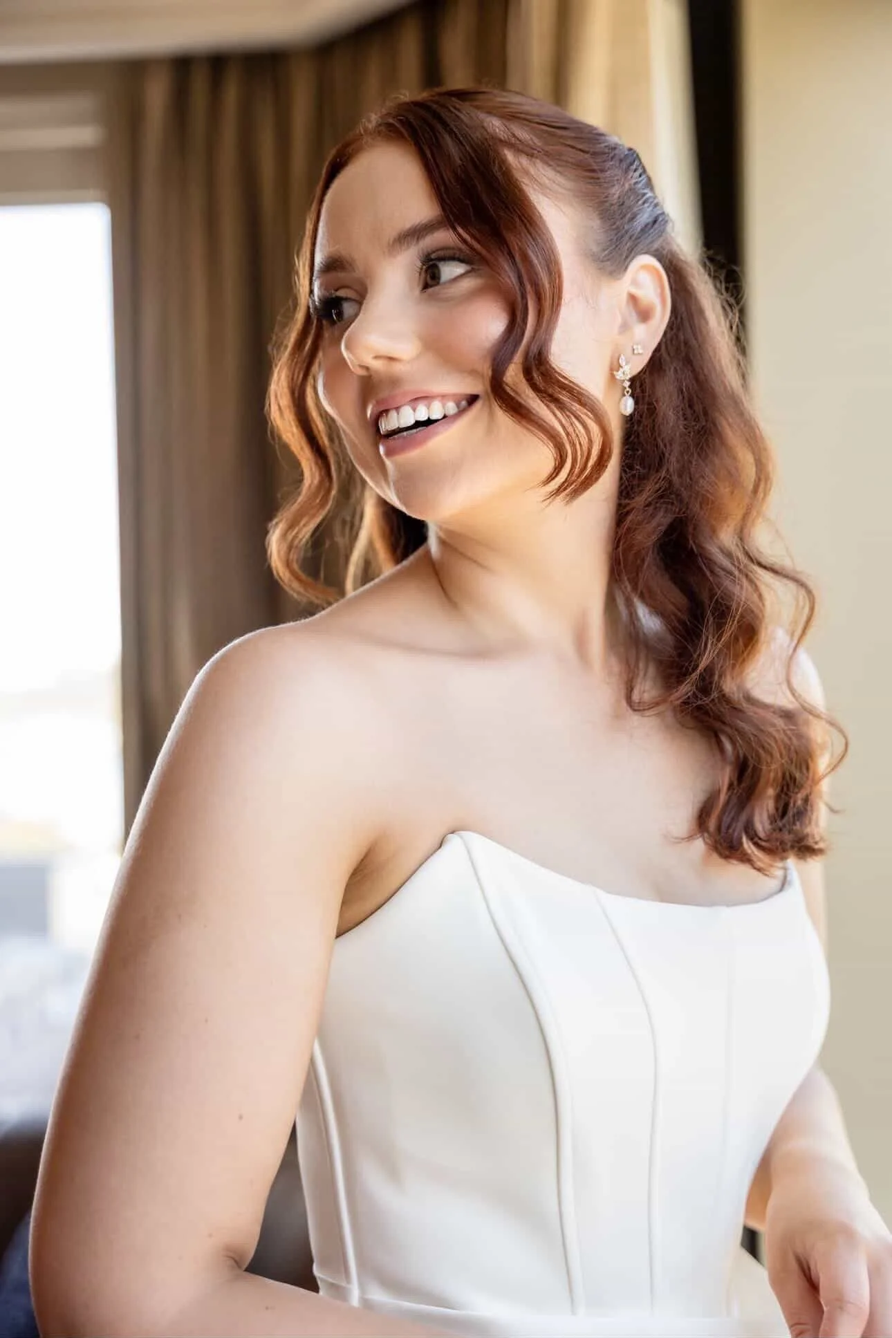 Close-up of a woman with red hair styled in loose waves, wearing earrings, smiling, dressed in a white strapless dress, standing in a room with curtains and a window in the background.