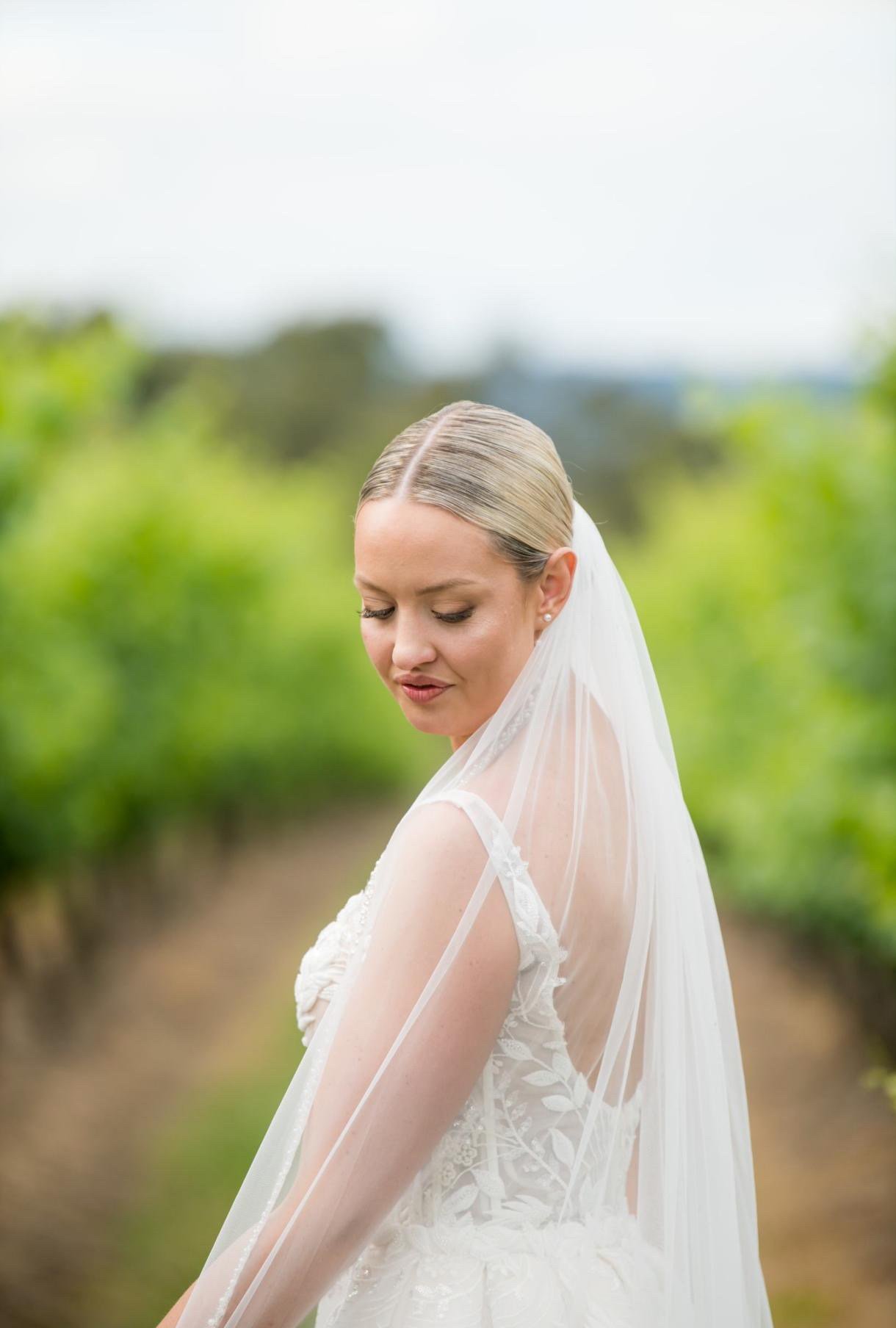 A bride in a wedding dress with lace details and a veil stands outdoors with greenery in the background, looking down with a serene expression.