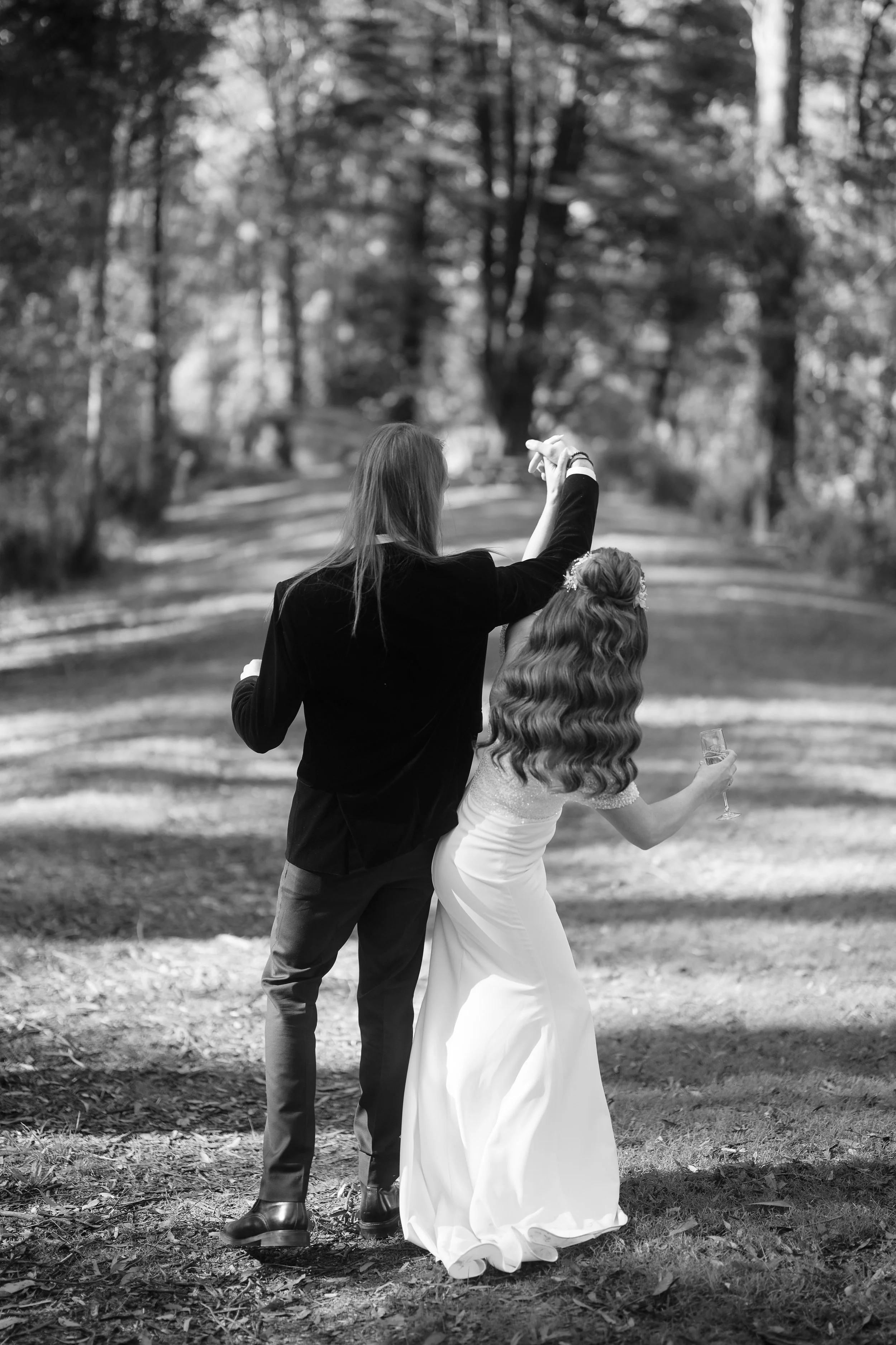 A couple dressed formally, the man in a suit and the woman in a wedding dress, walking together in a wooded area, holding hands with the woman holding a glass of champagne.