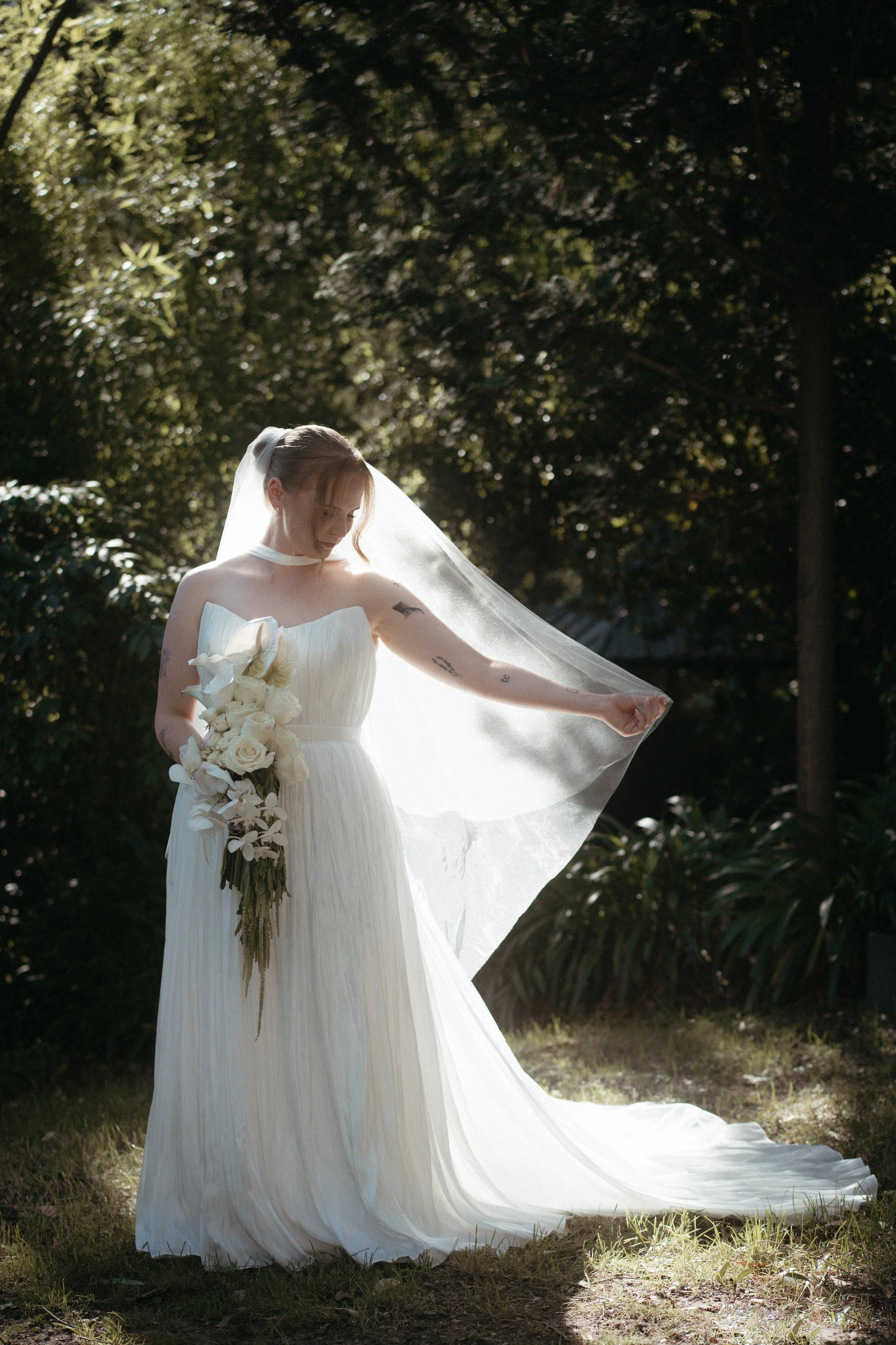 A bride in a white wedding gown holding a bouquet of white roses and calla lilies, standing outdoors with trees in the background, sunlight filtering through the leaves, and her veil flowing in the breeze.