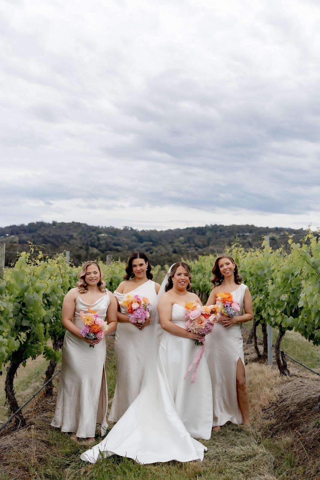 Four women in wedding dresses standing in a vineyard holding bouquets of pink, orange, and purple flowers.