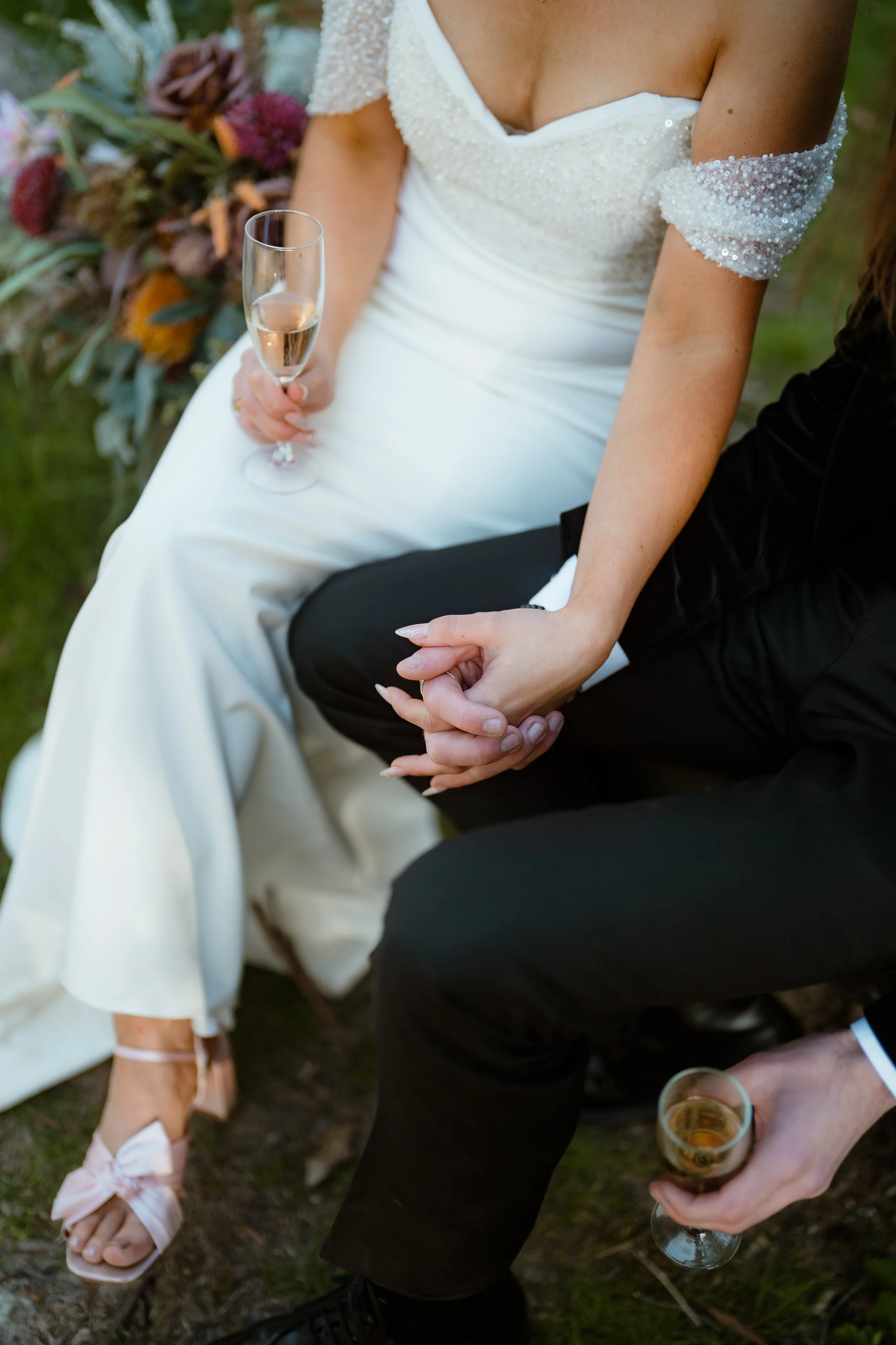 Close-up of a bride and groom holding hands at a wedding. The bride is in a white dress with beaded sleeves, and the groom is in a black suit. They are seated outdoors, with the bride holding a glass of champagne. In the background, a woman in a cream dress with pink strap heels is visible.