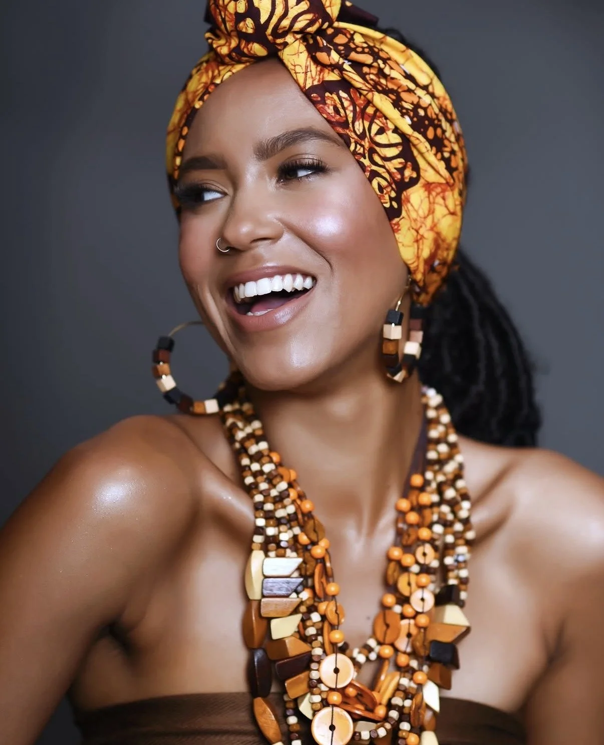 A woman with a wide smile, wearing a colorful patterned head wrap and large beaded jewelry, including necklaces, earrings, and a nose ring, against a dark background.