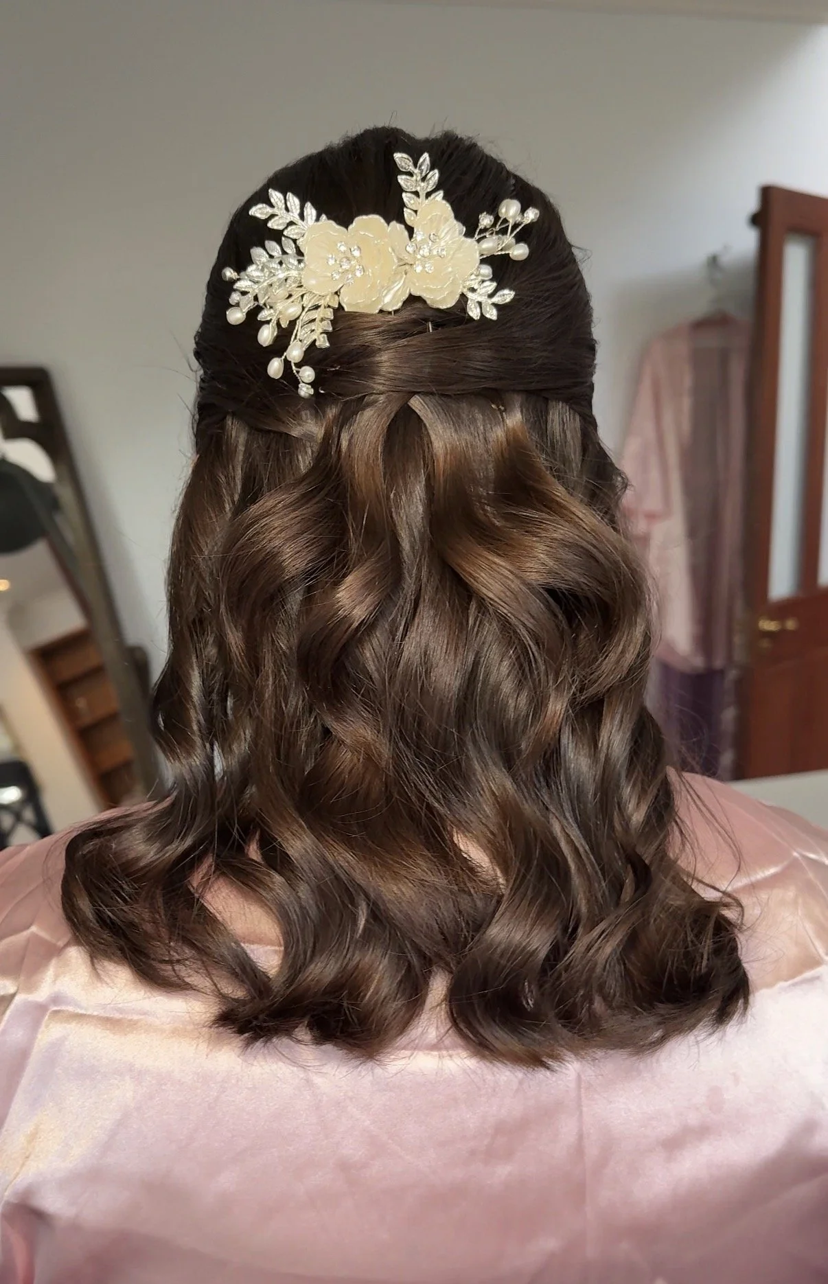 Back view of a woman with long, wavy brown hair styled in loose curls, wearing a floral hair accessory with pearls and white flowers. The woman is sitting on a pink surface with a background featuring a mirror, wooden furniture, and a light-colored w