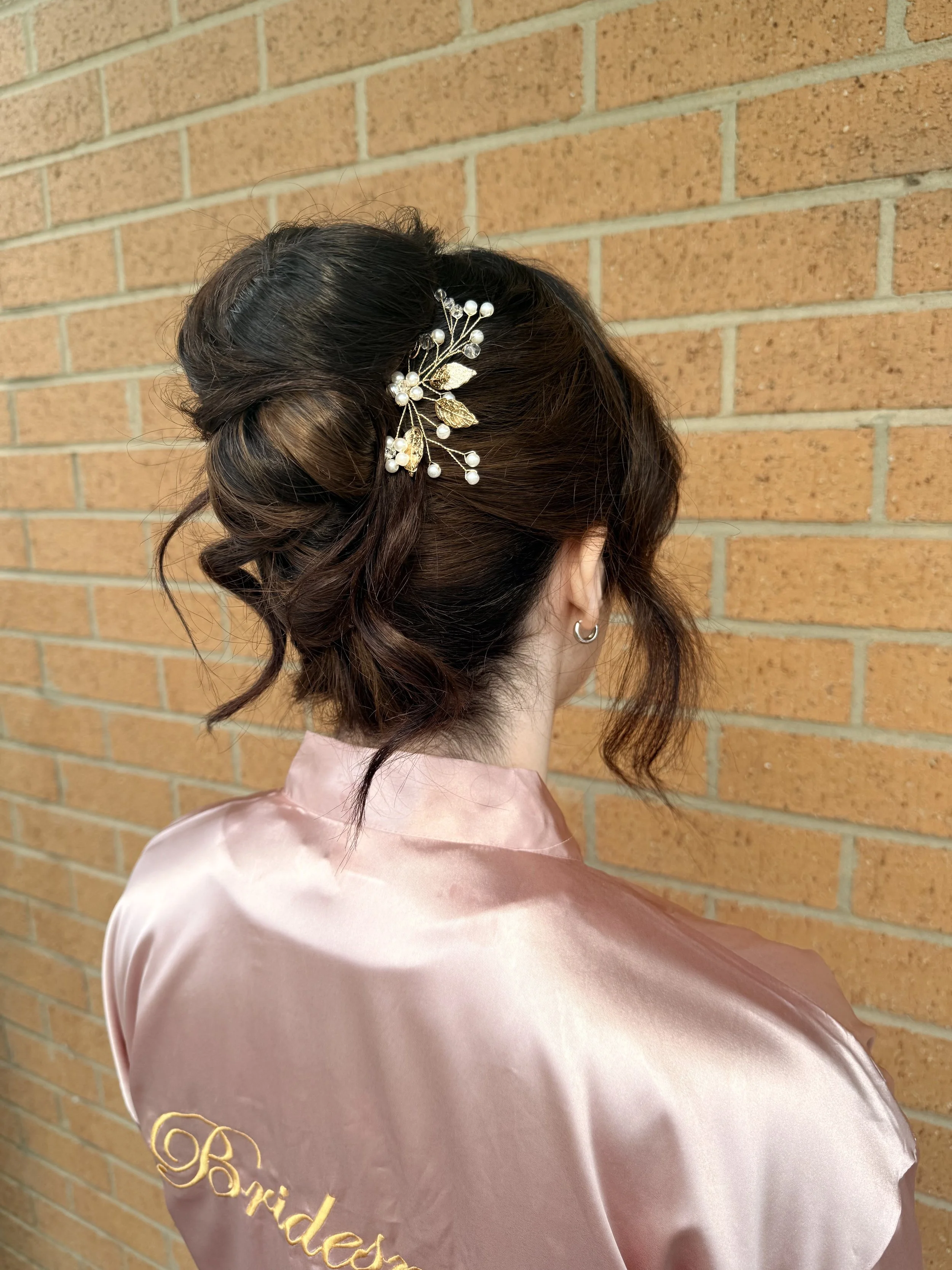 Side view of a woman with styled brown hair adorned with an elegant hairpin featuring white pearls and gold leaves, wearing a pink satin robe with embroidered text, standing against a brick wall.