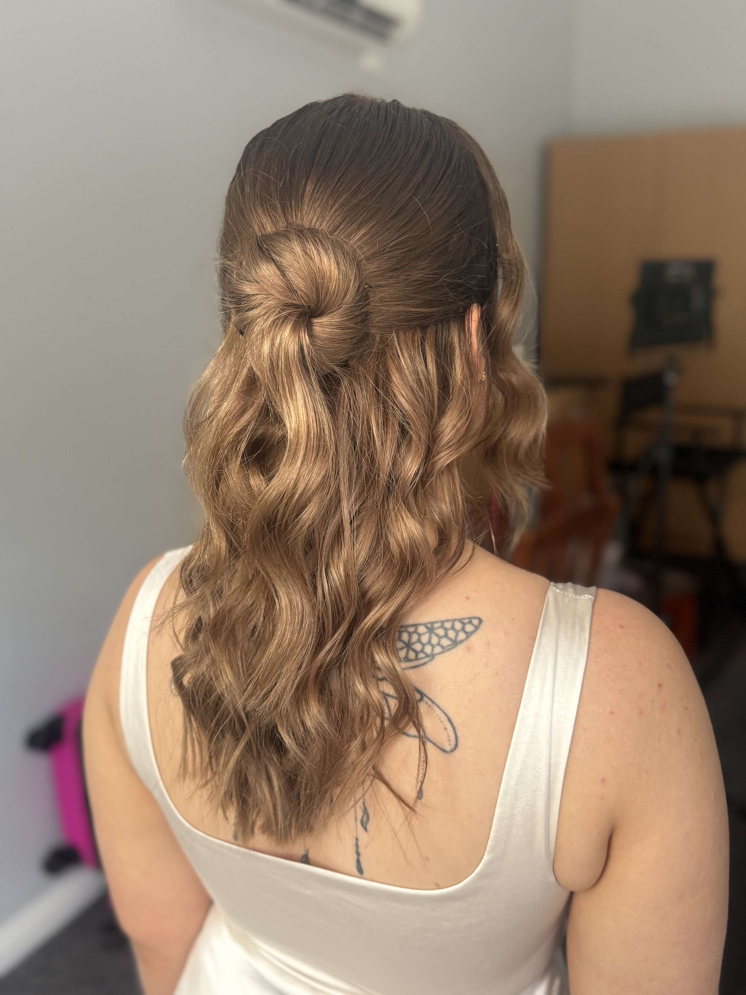 A woman with wavy, reddish-brown hair styled in a loose bun with curly strands hanging down, wearing a white tank top and a tattoo of a leaf on her shoulder, in a room with a gray wall and a piece of furniture in the background.