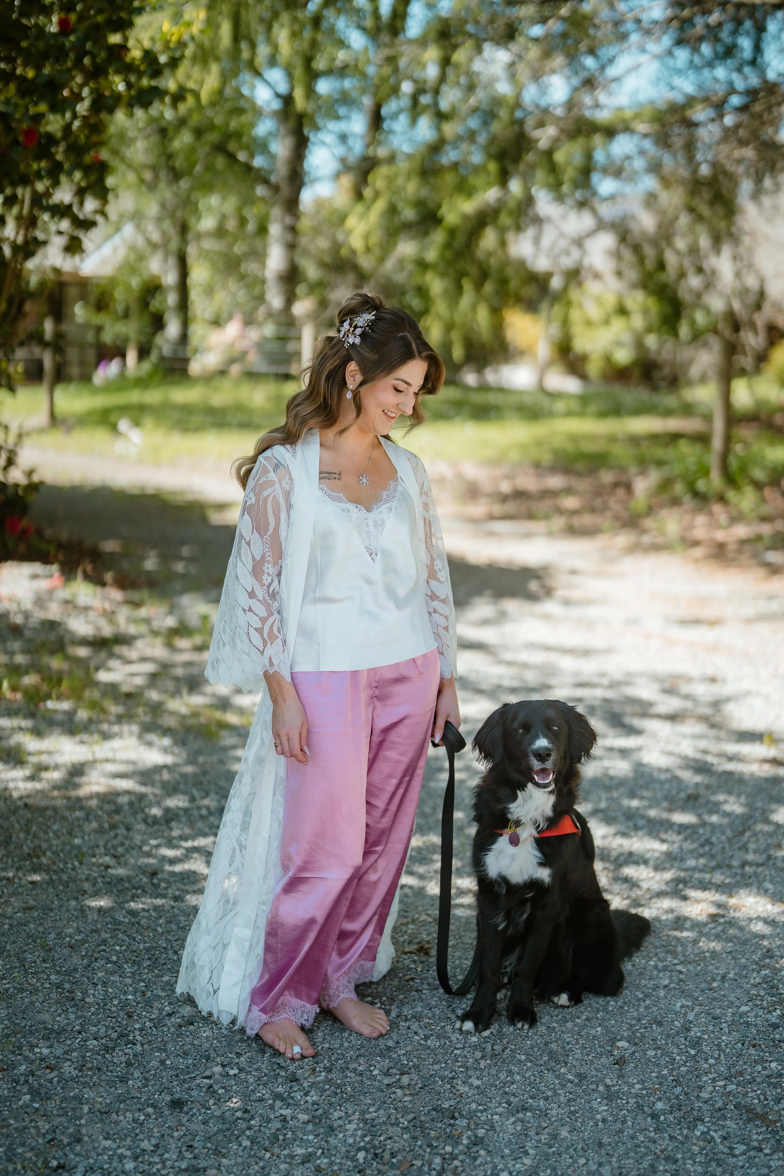 Woman in pink satin pants and white lace top, barefoot, standing outdoors with black and white dog on leash, smiling, in a sunny park with green trees.