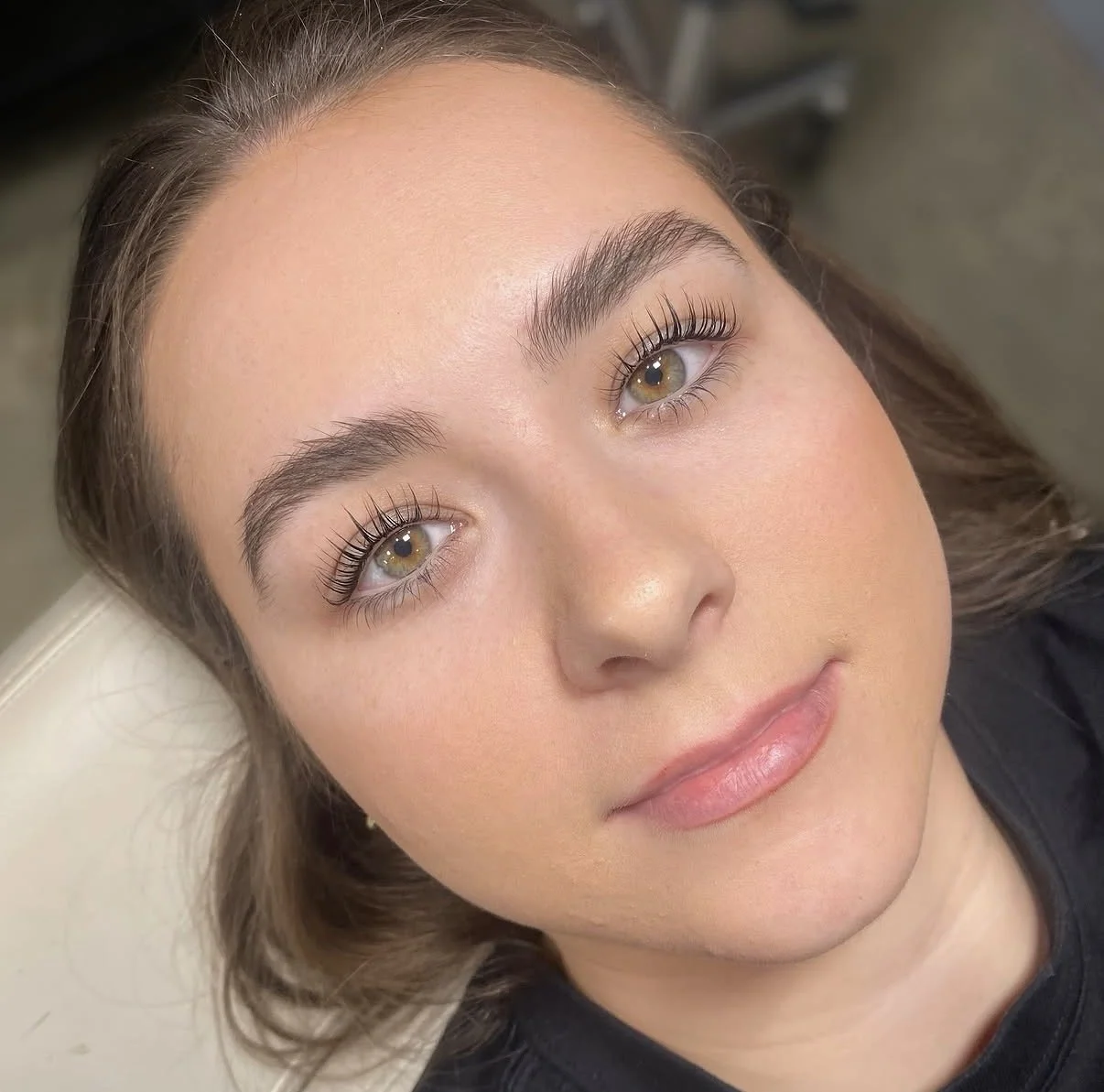 Close-up of a young woman's face with clear skin, hazel eyes, and well-groomed eyebrows, lying down with her head tilted slightly, looking directly at the camera.