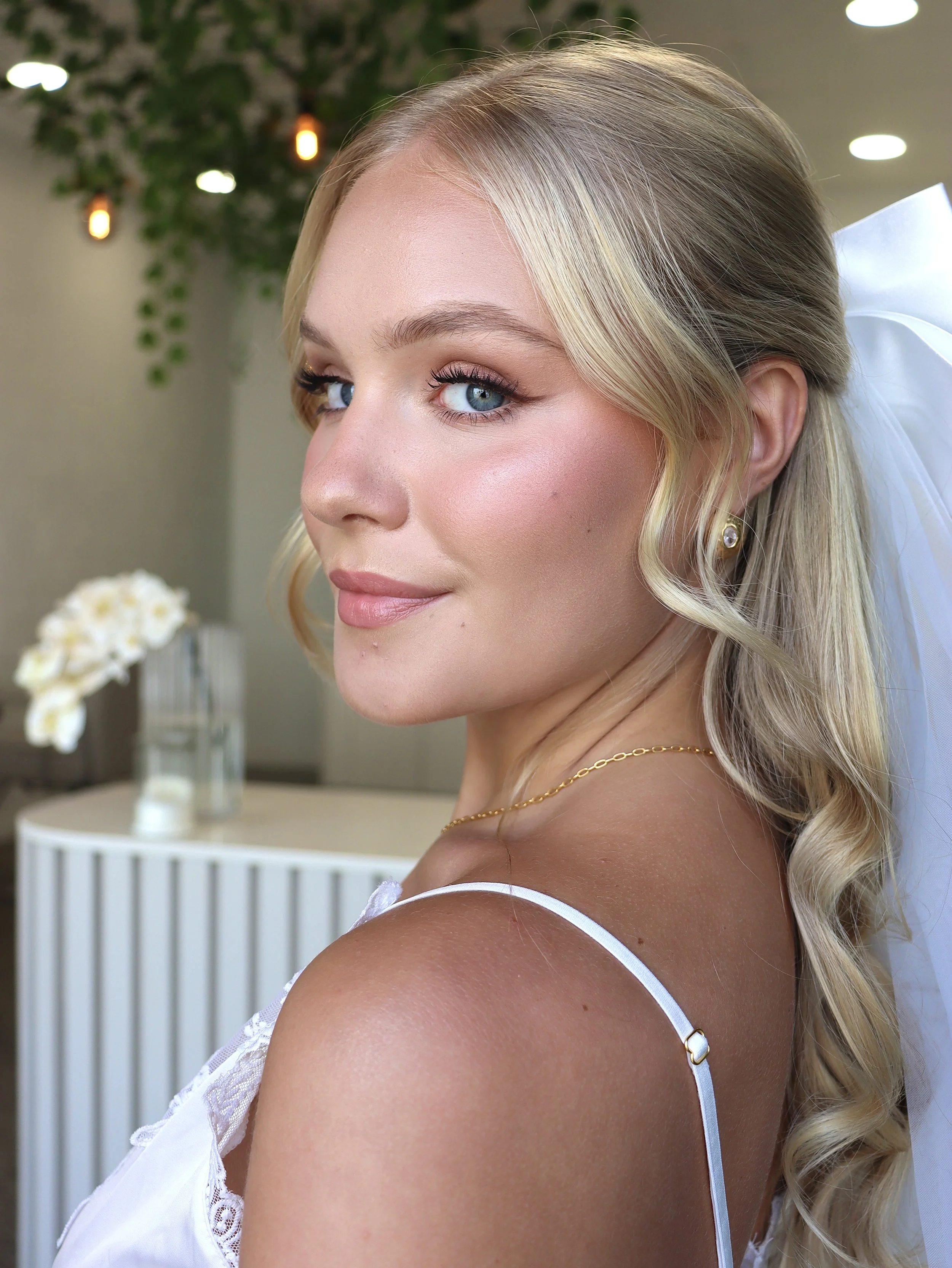 Close-up of a smiling woman with blonde hair, blue eyes, wearing pearl earrings and a white dress, with a background of green plants, hanging lights, and white flowers.