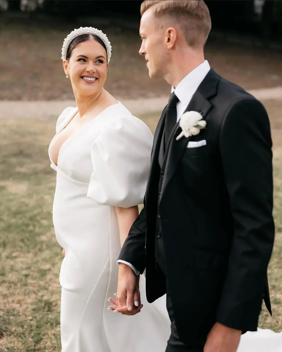 A bride and groom holding hands and walking outside on their wedding day. The bride is wearing a white wedding dress and the groom is in a black suit with a white boutonniere.
