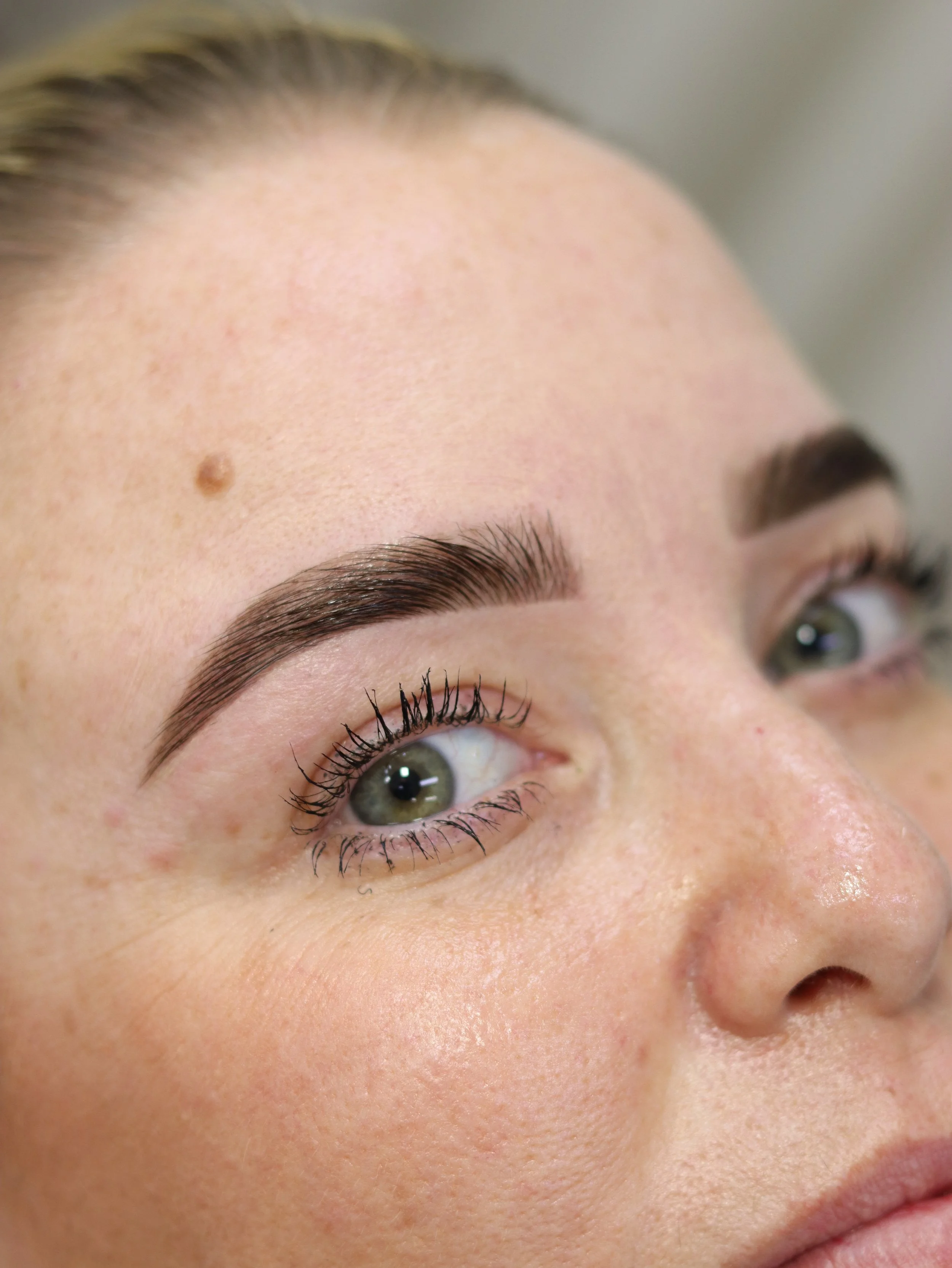 Close-up of a woman's face, showing her eye, part of her eyebrow, and forehead with a mole, and her lips.