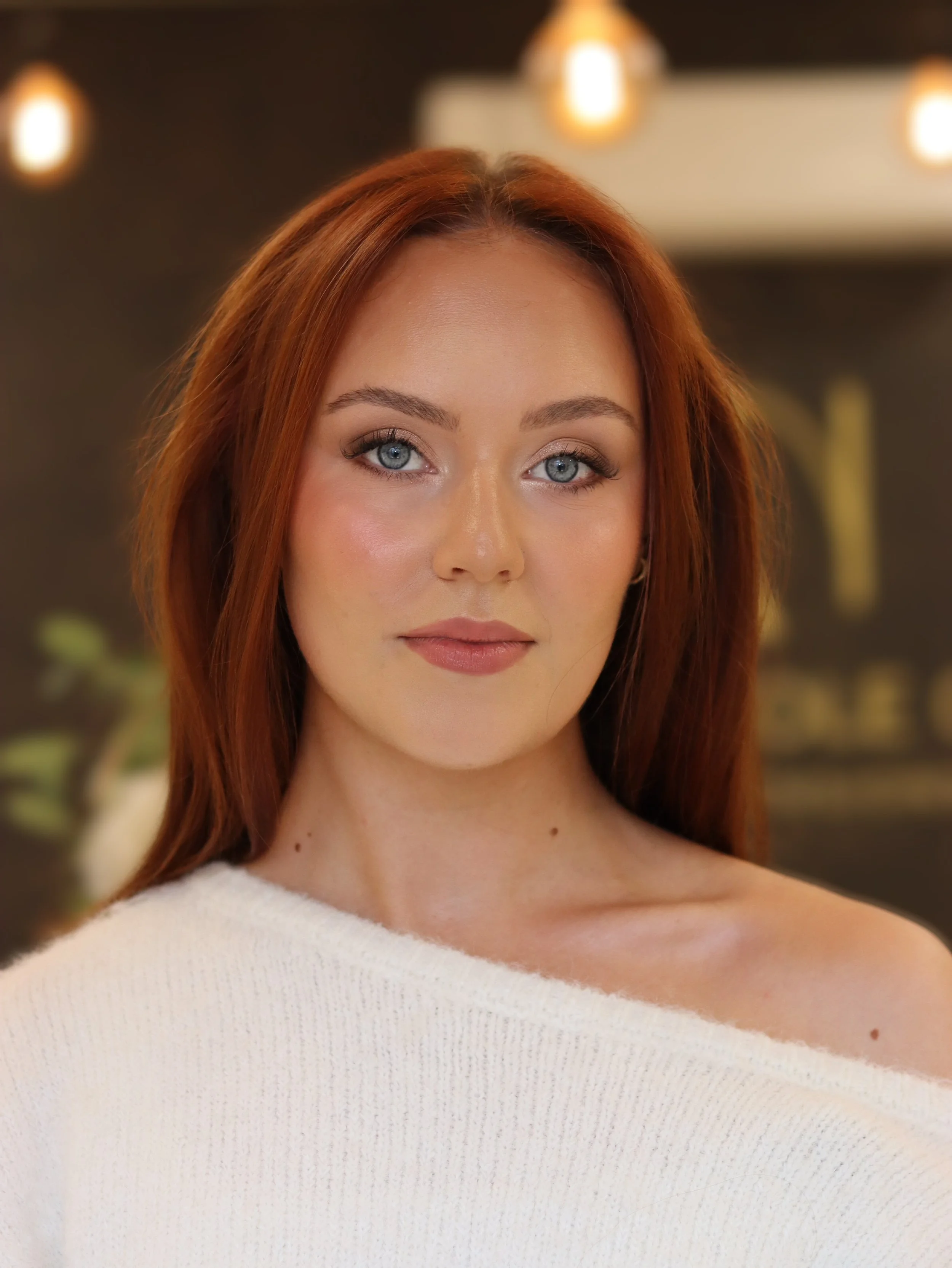 Close-up of a woman with red hair, blue eyes, and light makeup, wearing a white off-the-shoulder sweater, in an indoor setting with warm lighting.