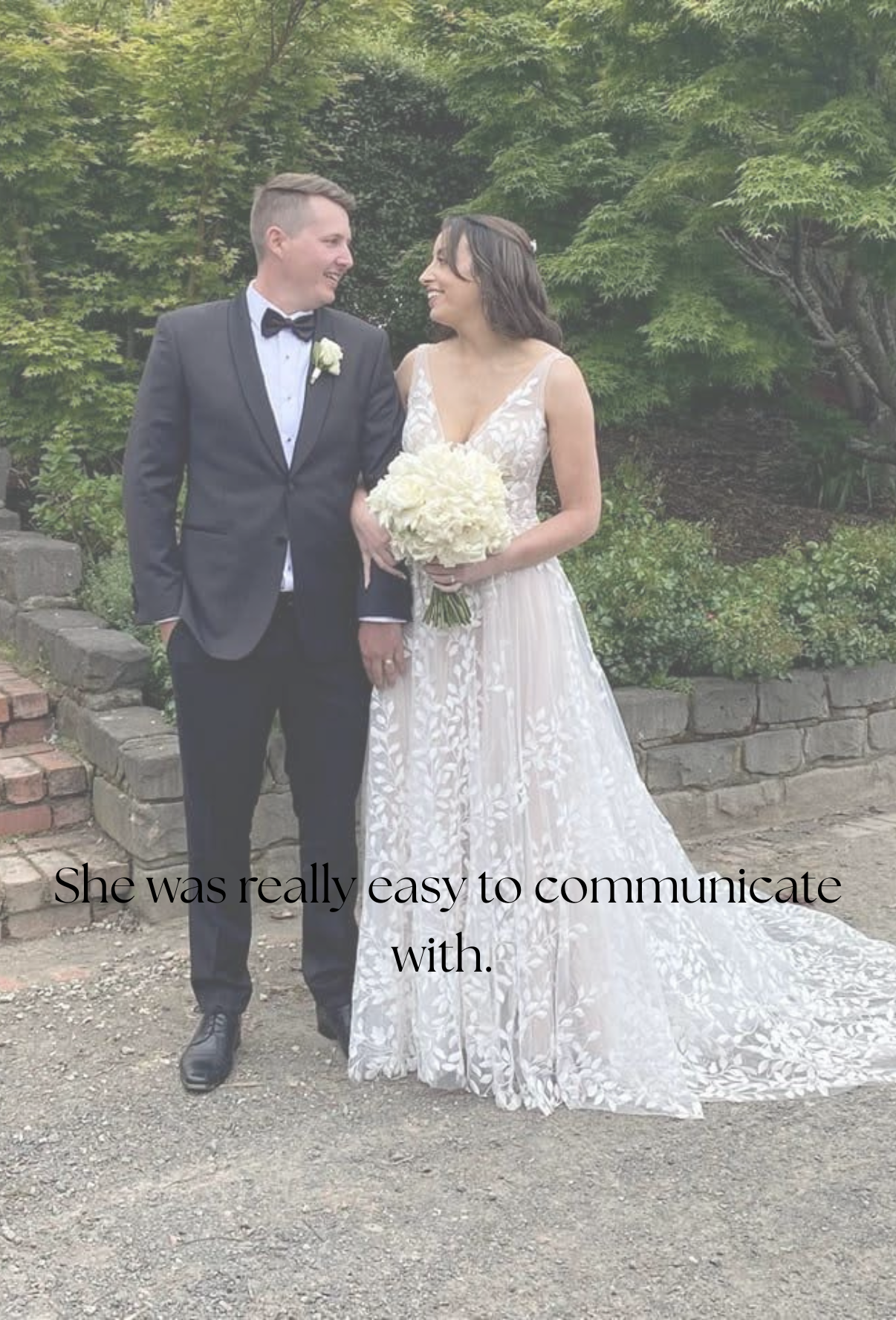 A newlywed couple standing outdoors on a garden path, holding hands and smiling at each other. The groom is wearing a black tuxedo with a bow tie, and the bride is wearing a white lace wedding gown and holding a bouquet of white flowers. Green trees and shrubs are in the background.
