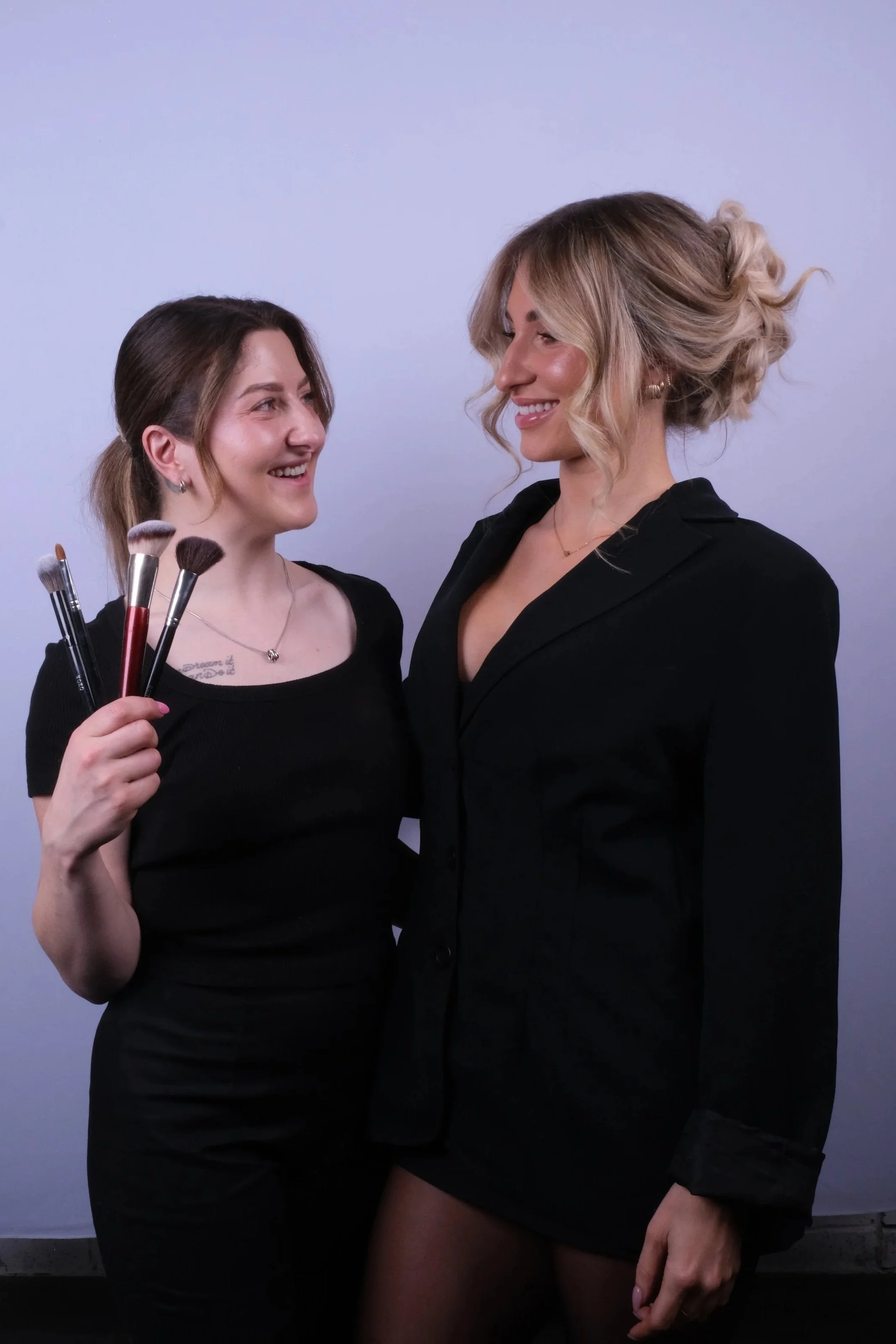 Two women standing close together, smiling at each other, with one holding makeup brushes.