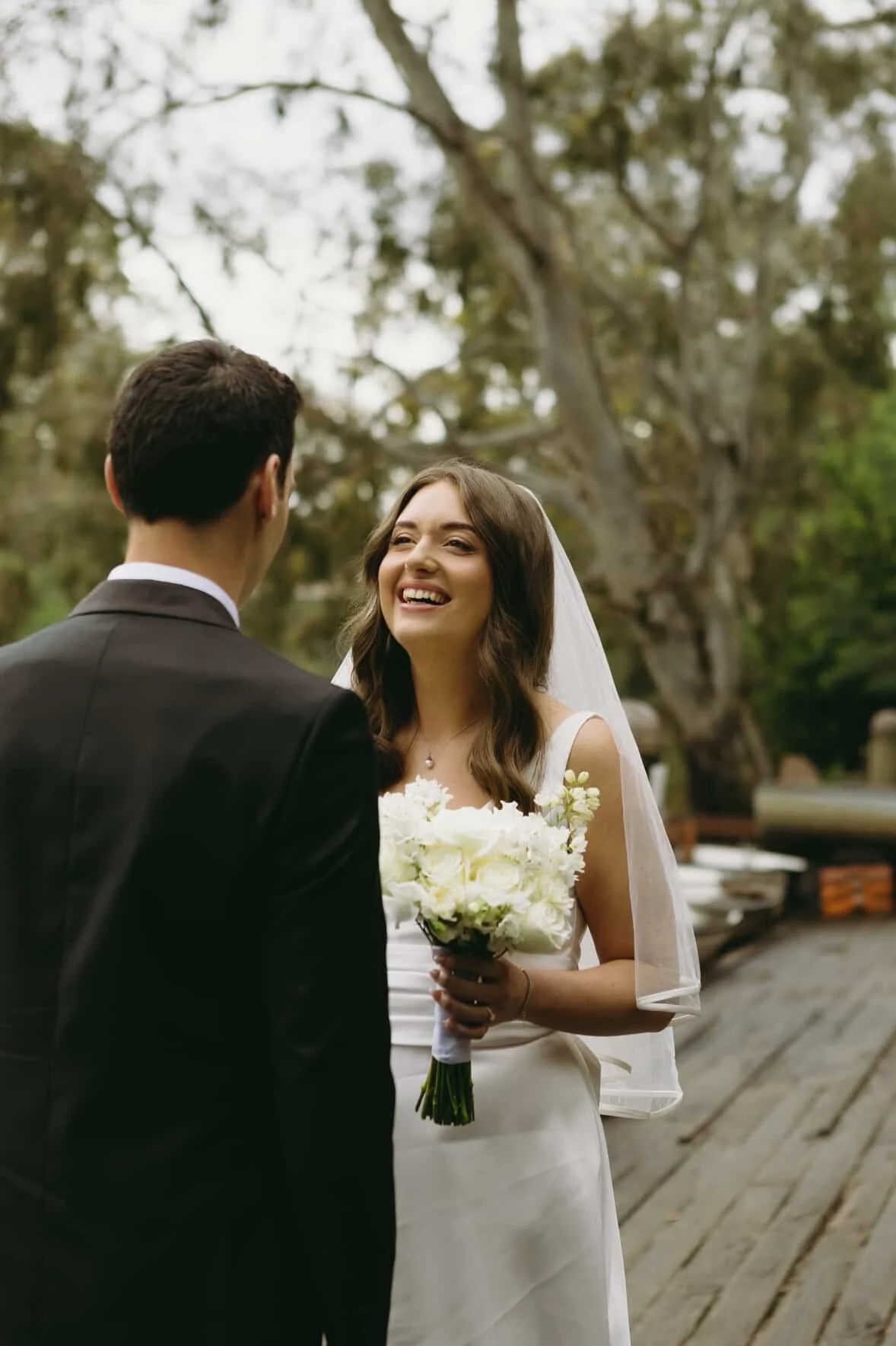 A bride and groom on their wedding day outdoors, with the bride smiling and holding a bouquet of white flowers, wearing a white dress and veil, and the groom wearing a black suit, in front of a background of trees.