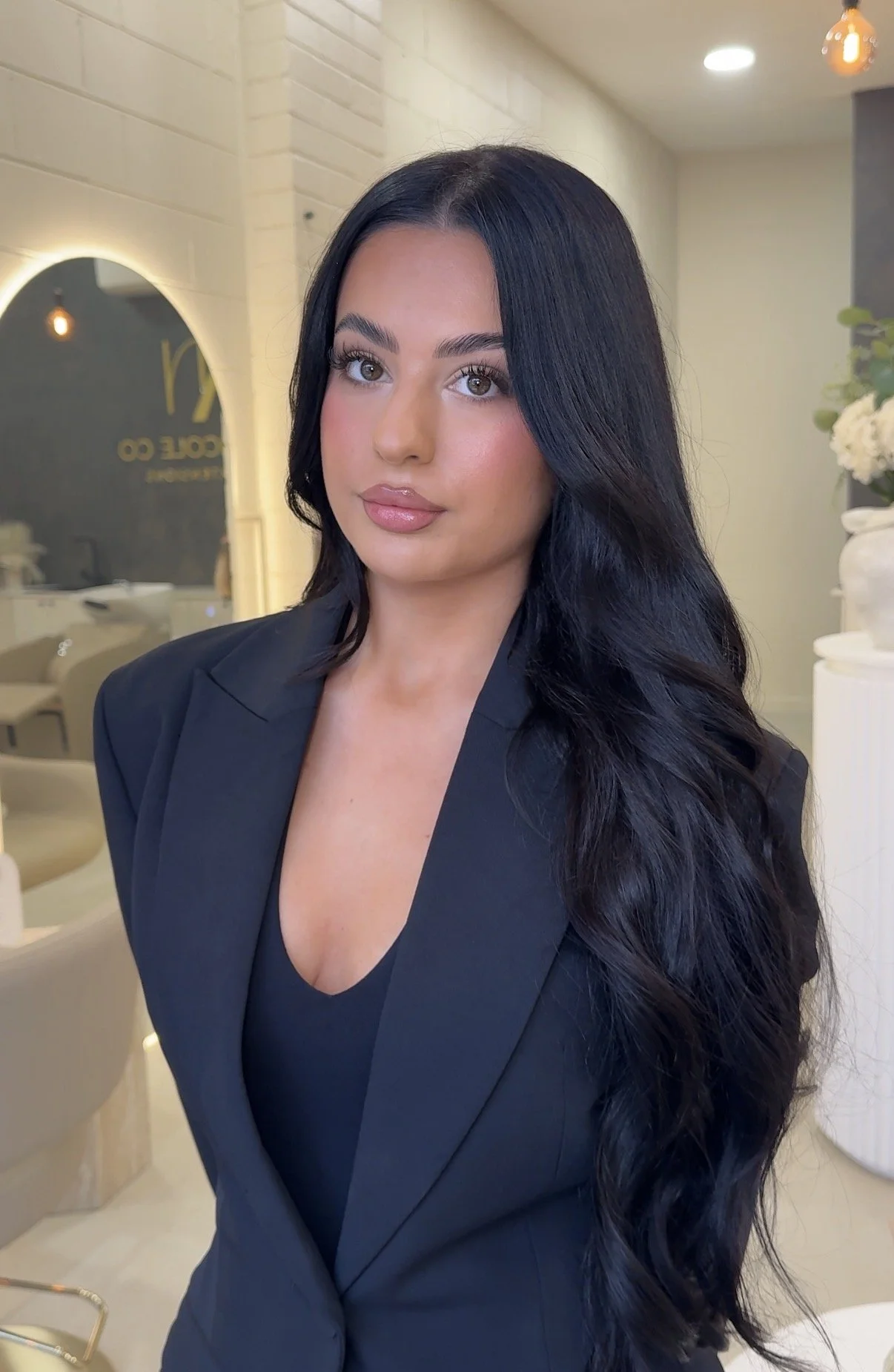 A woman with long black hair and fair skin wearing a dark blazer and top in a modern, well-lit interior space.