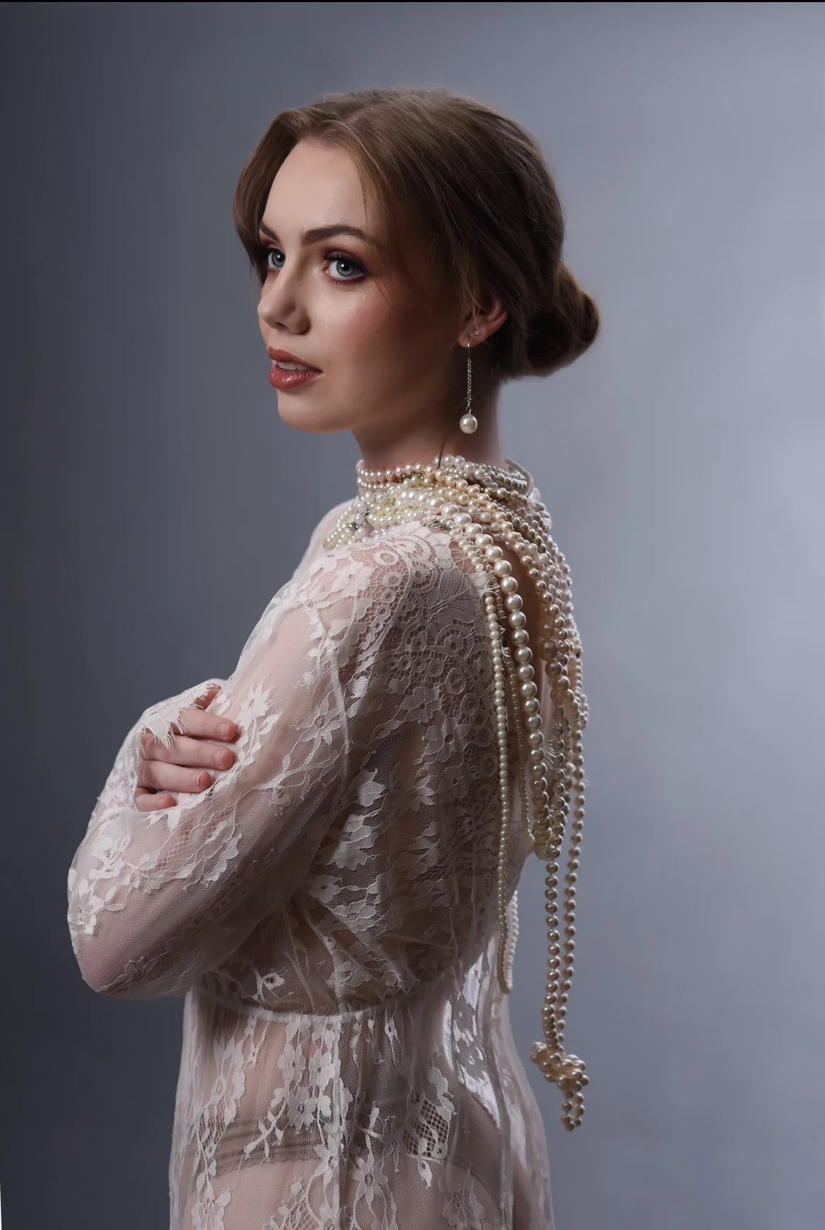 A young woman with short brown hair styled in a low bun, wearing a lace dress and multiple strands of pearls around her neck, with one earring visible, posing against a plain gray background.