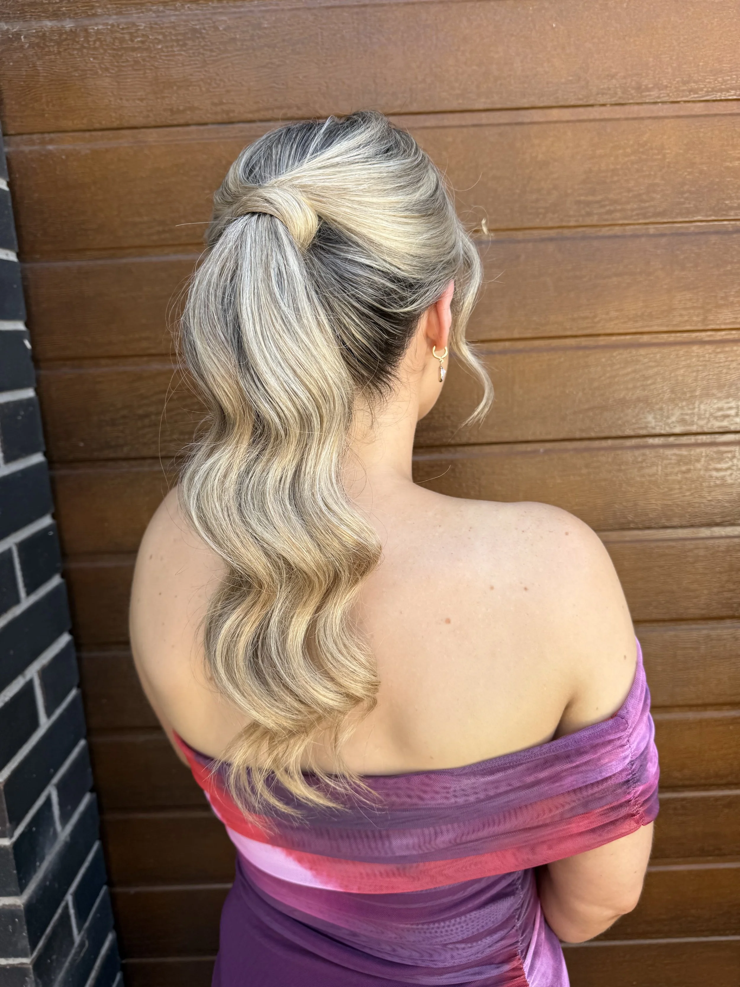 Back view of a woman with wavy blonde hair styled in a loose ponytail, wearing a purple off-the-shoulder top, standing against a wooden wall.