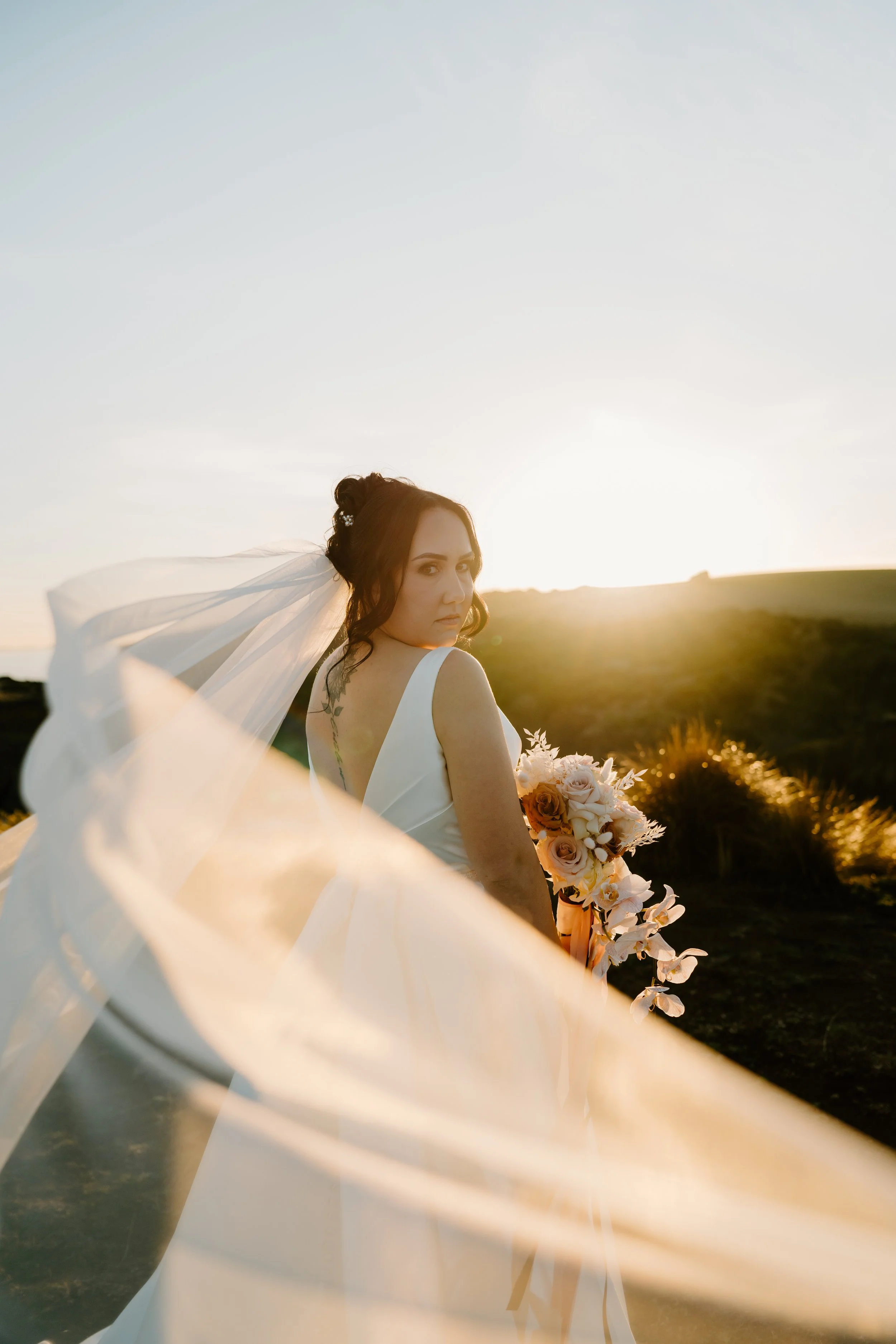 A woman in a white wedding dress holding a bouquet of flowers, standing outdoors at sunset, with a flowing veil and lush green landscape in the background.