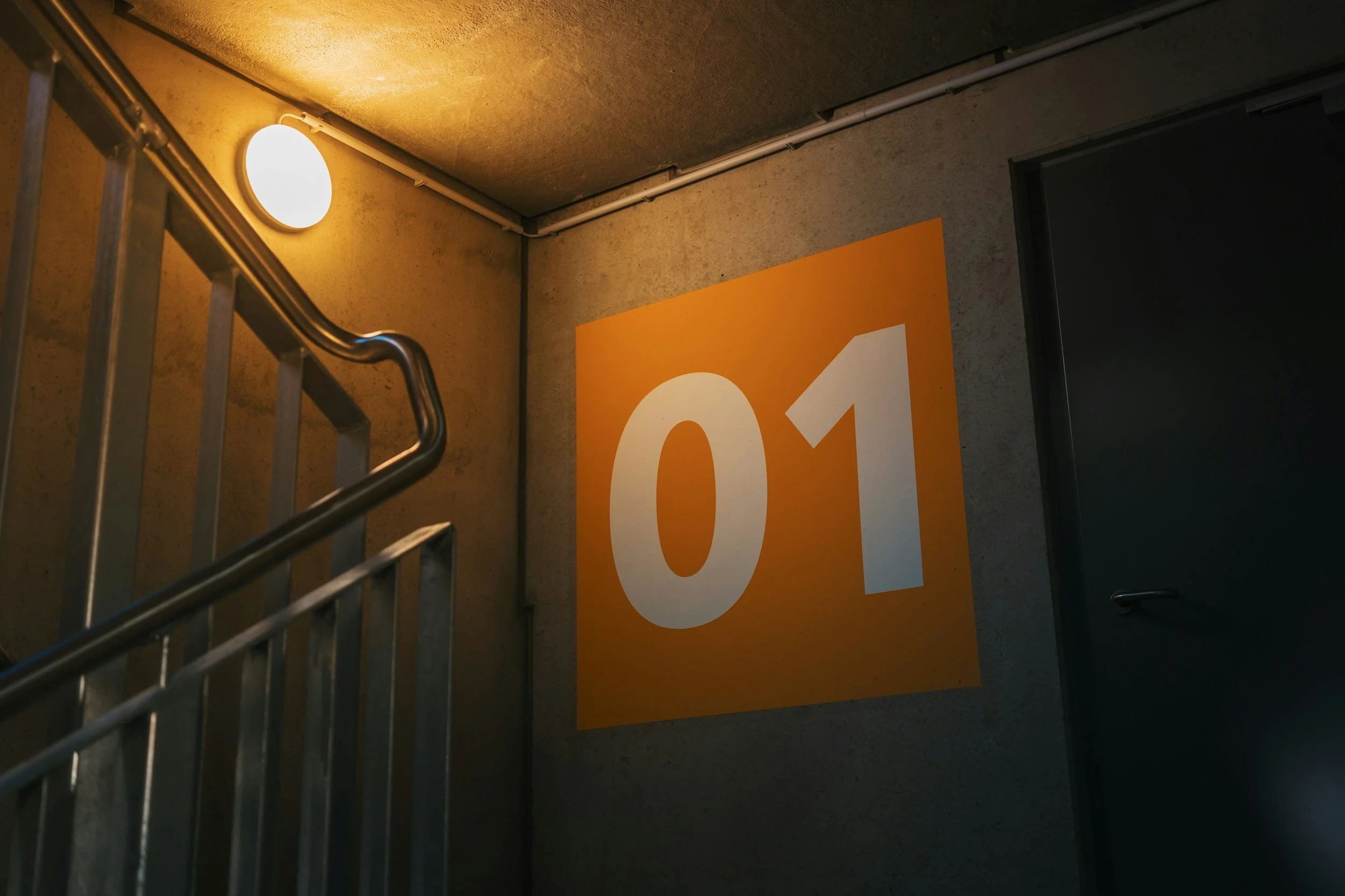 Indoor staircase with a large orange sign displaying the number 04, a wall-mounted light, and visible piping along the ceiling.