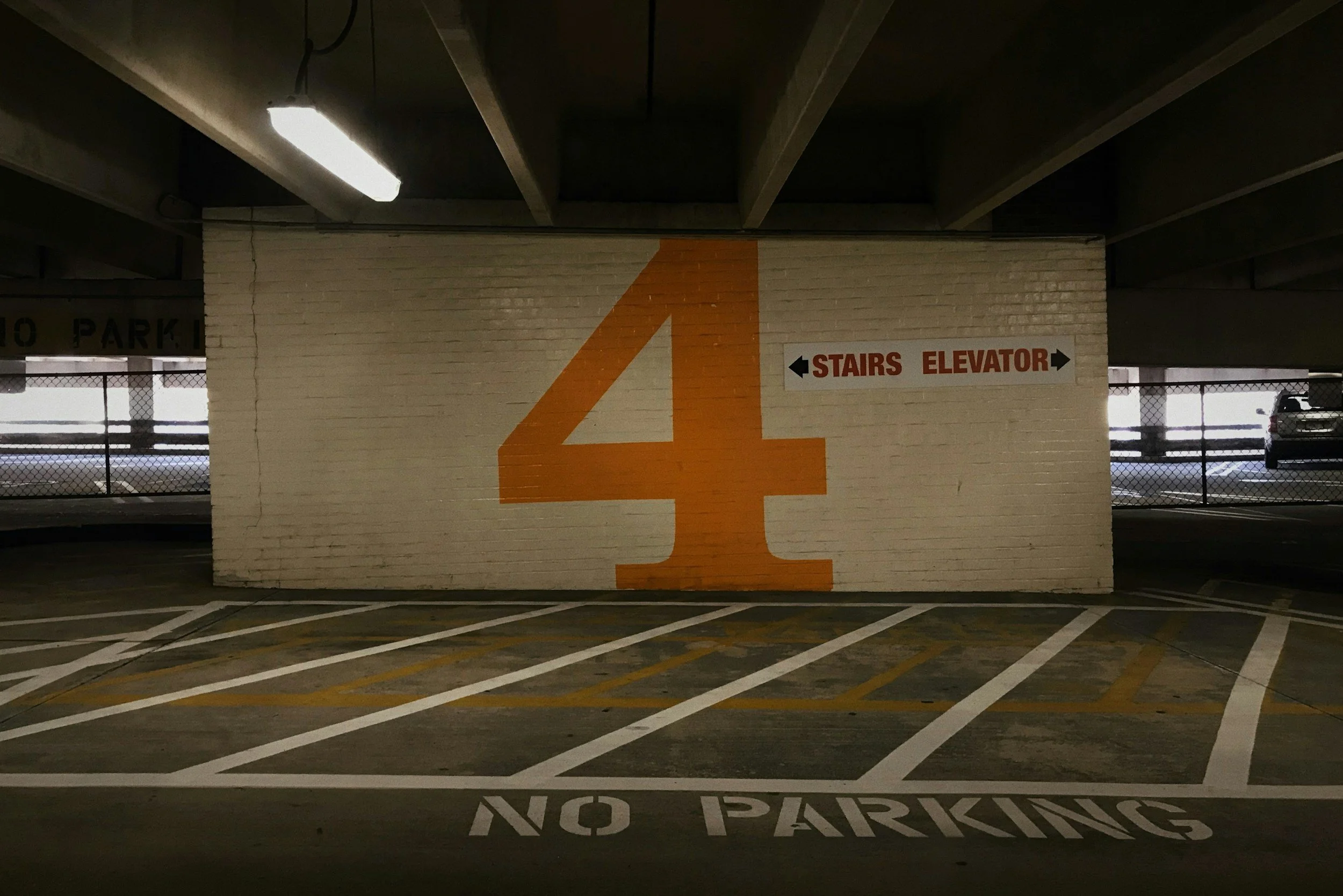Parking garage with a large orange number 4 painted on a white brick wall. Sign on the wall indicates stairs and elevator accessible in both directions. Parking spots marked with white lines on the ground, and the area is dimly lit with ceiling lights.