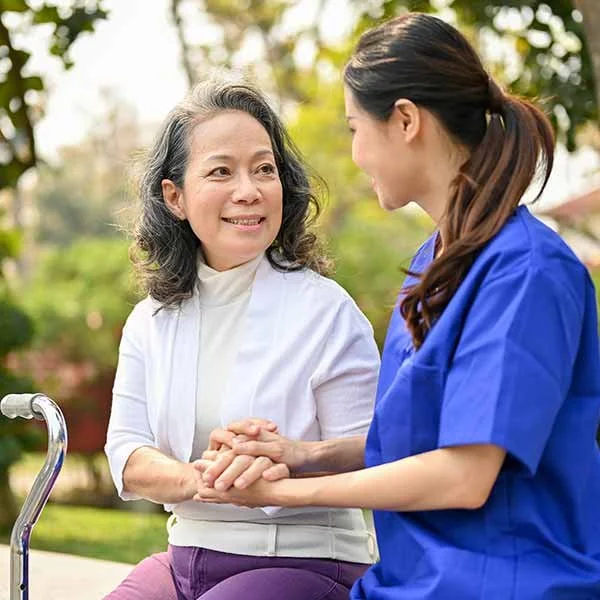 An elderly woman in a white jacket sitting outdoors and holding hands with a young female caregiver in blue scrubs, both smiling at each other amidst a background of greenery.