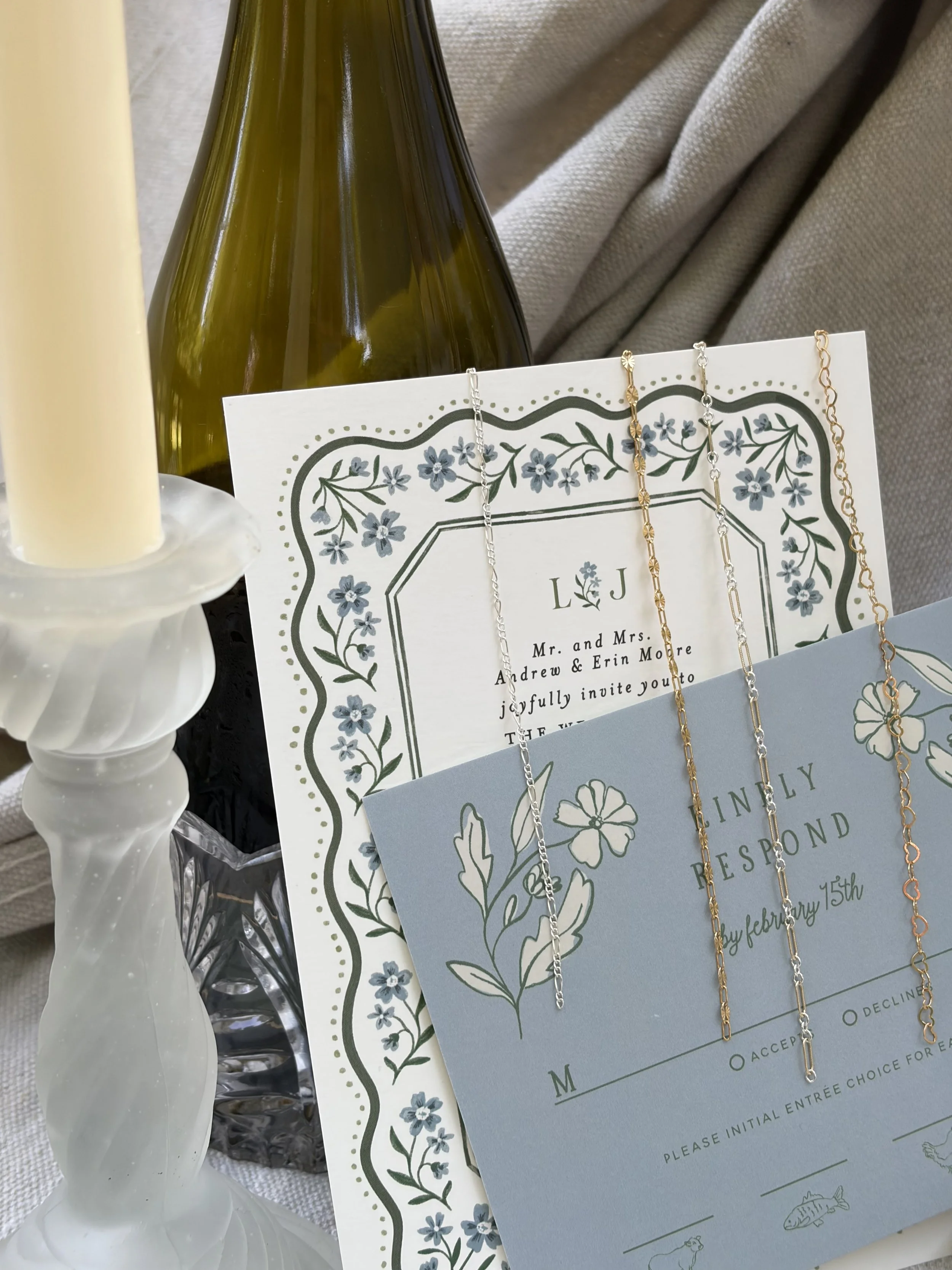 A wedding invitation with a blue floral border and the text 'Mr. and Mrs. Andrew & Erin Moore invite you to joyfully invite you to the wedding.' There are three necklaces hanging on the invitation, and a green glass bottle behind it. To the left, there is a white candlestick with a cream-colored candle, and part of a patterned linen cloth is visible.