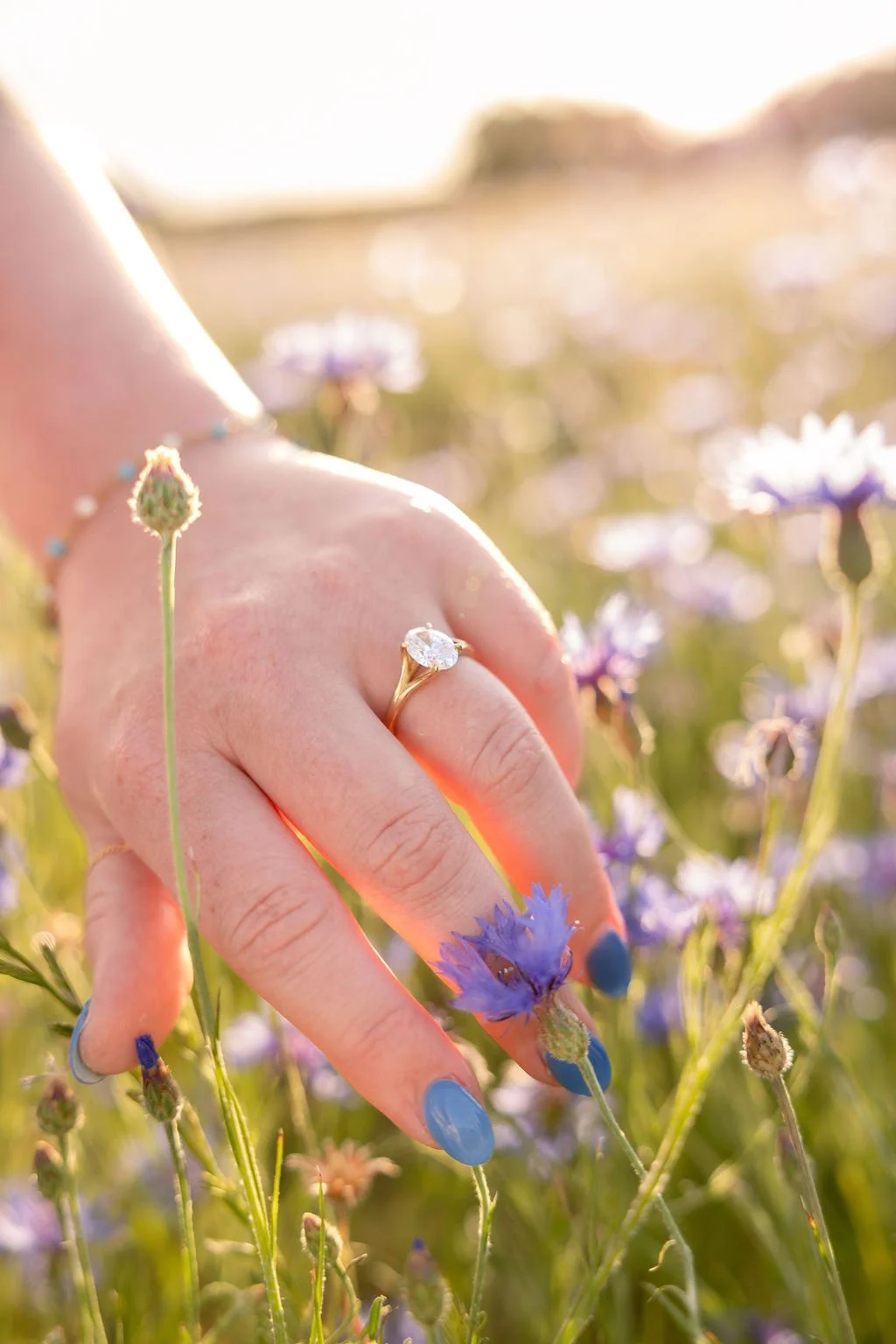 Close-up of a person's hand with a diamond engagement ring, surrounded by purple field flowers during sunset.